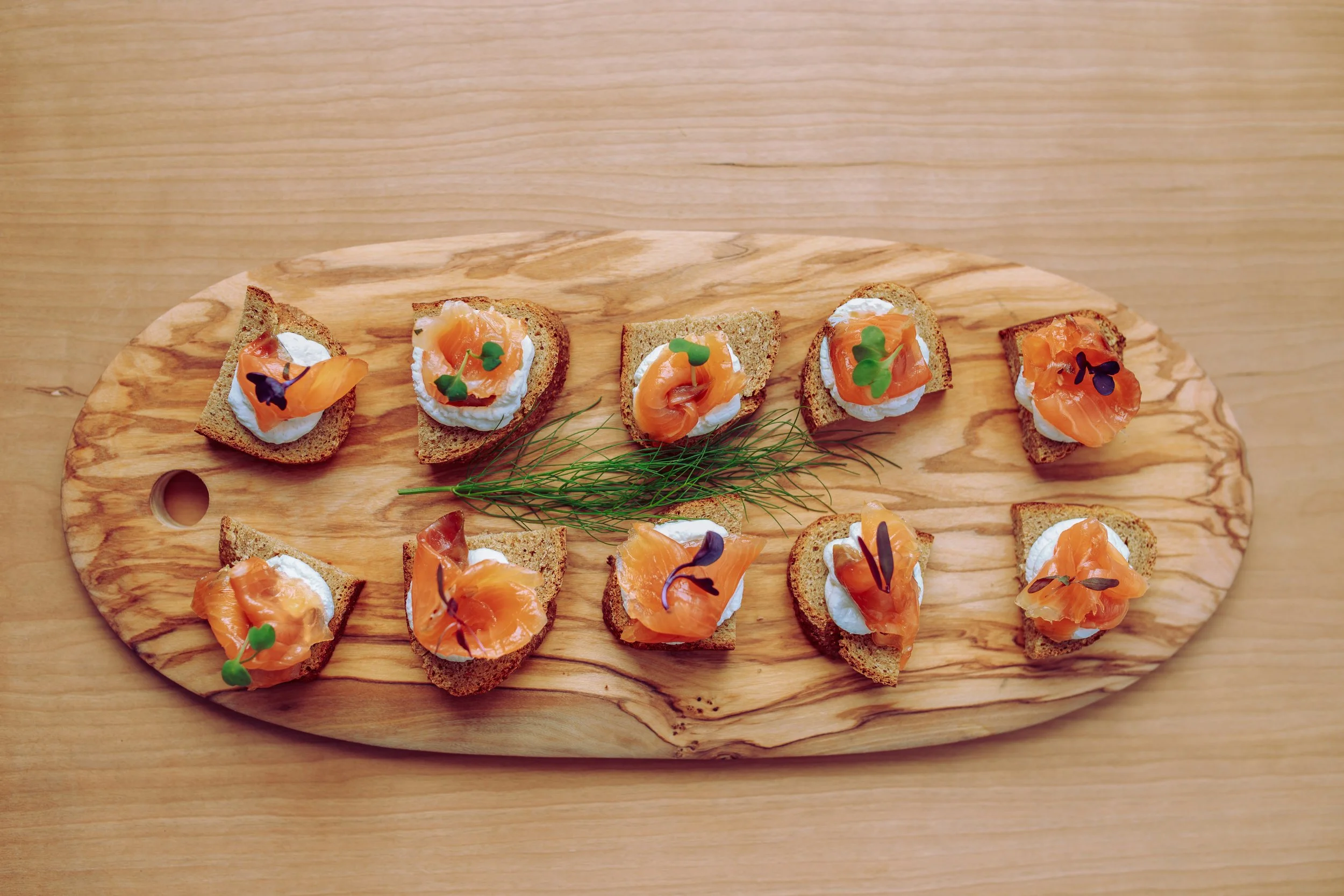 Several slices of bread topped with cream cheese, smoked salmon, and microgreens on a wooden serving board.