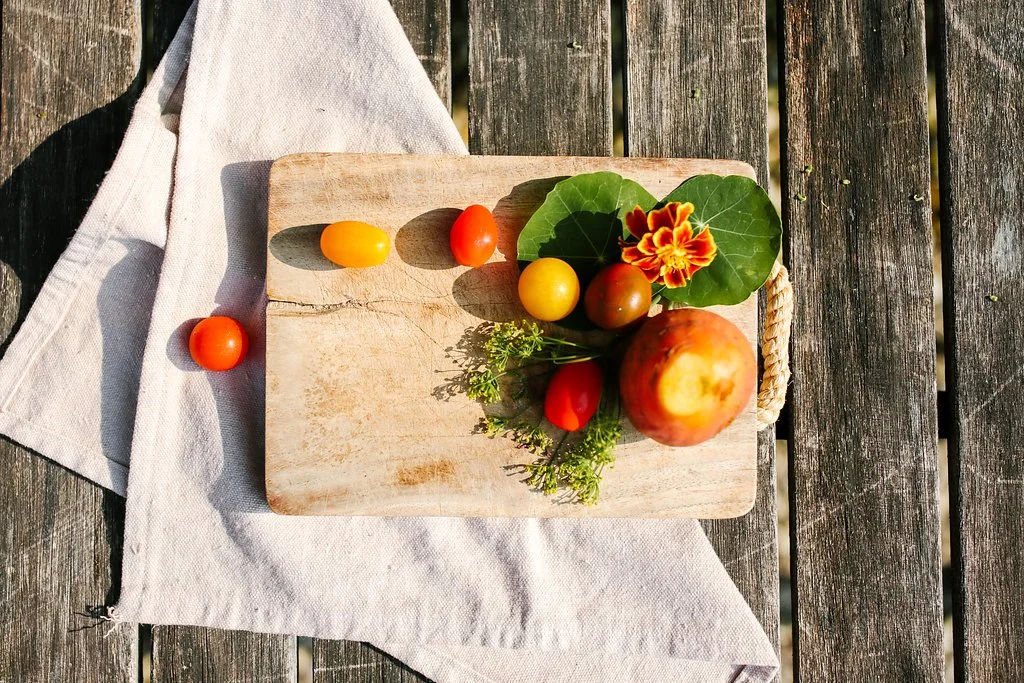 A small wooden cutting board with various tomatoes, a large apple, green leaves, colorful flowers, and fresh herbs on a white cloth outside on a wooden surface.