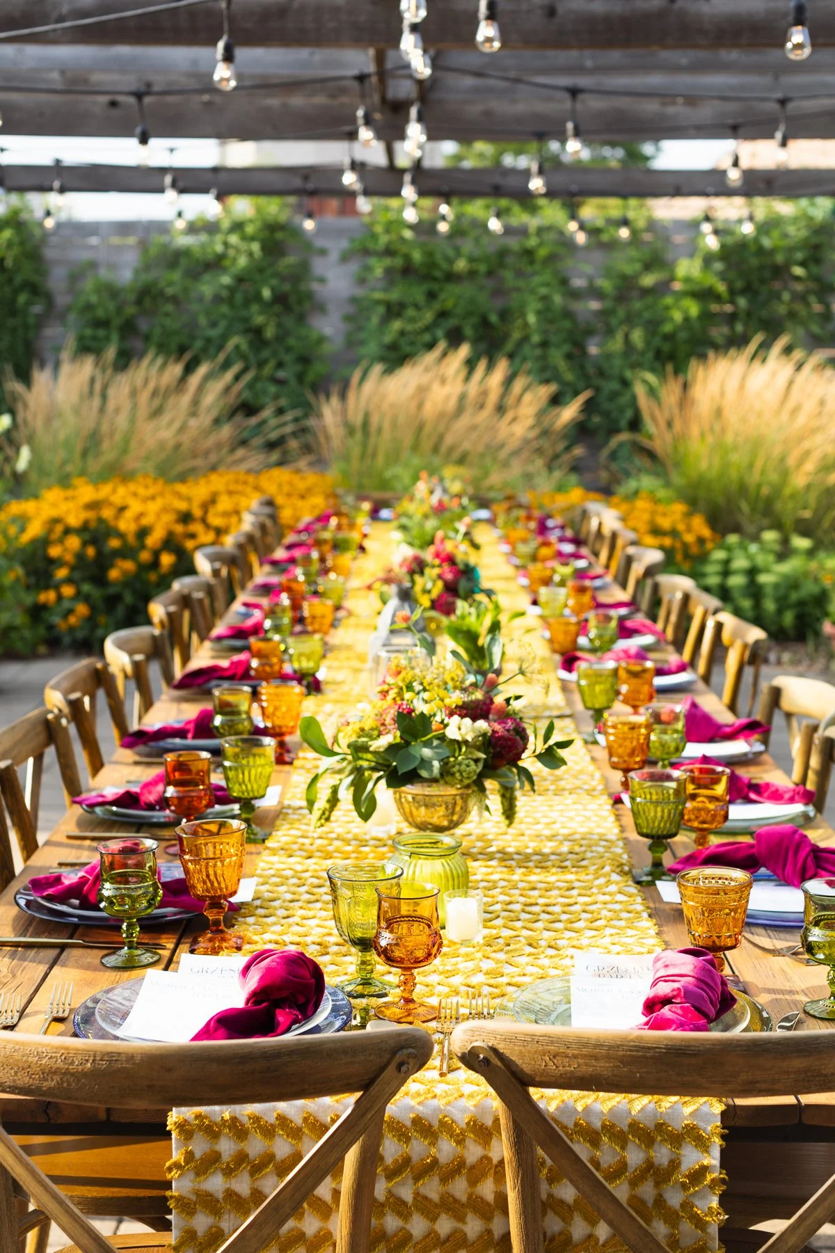 A long outdoor dining table decorated with floral centerpieces, colorful glassware, and pink napkins, set in a garden with string lights overhead and greenery in the background.