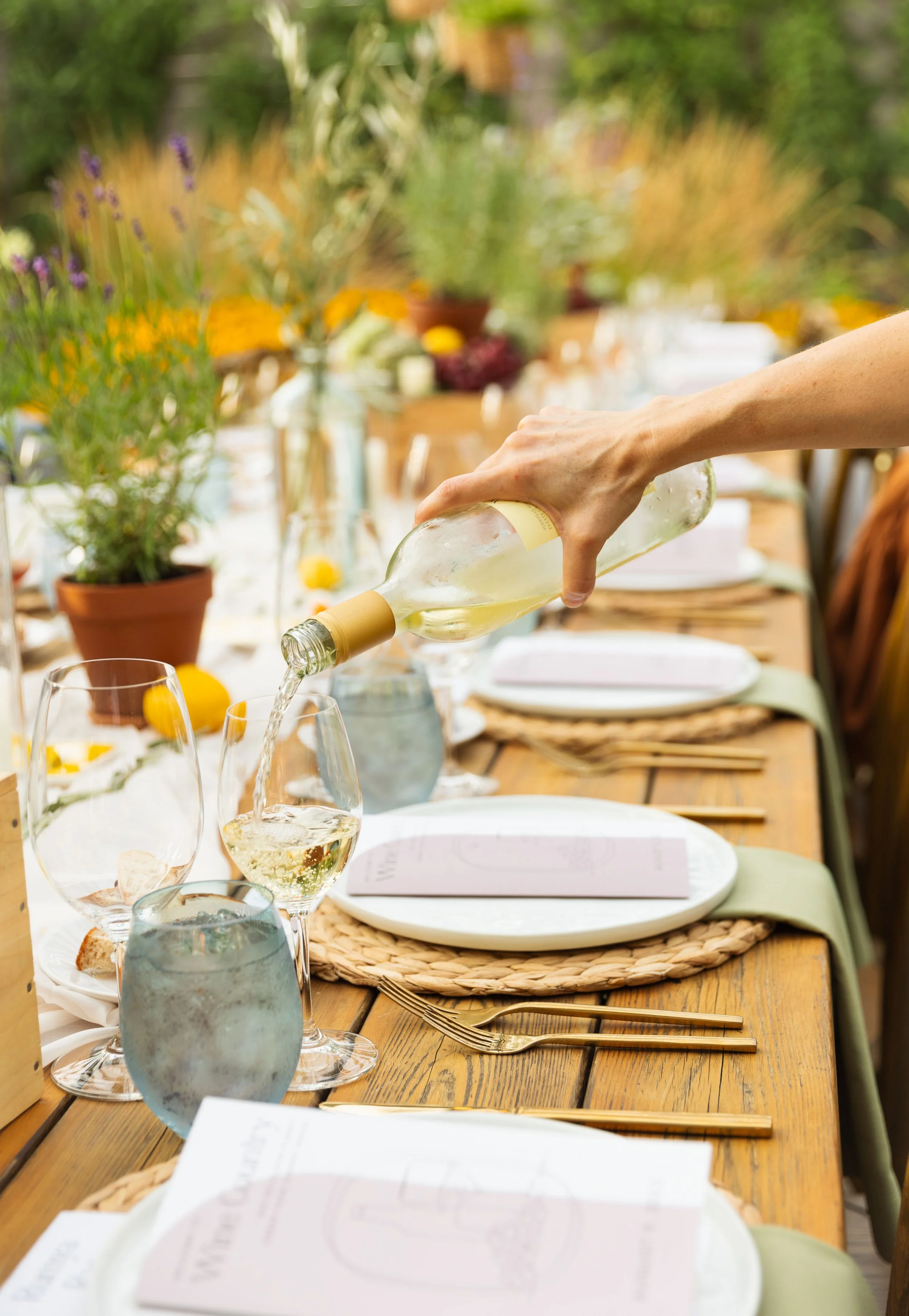 A person pouring white wine into glasses on a wooden dining table set outdoors with flowers, potted plants, and place settings.