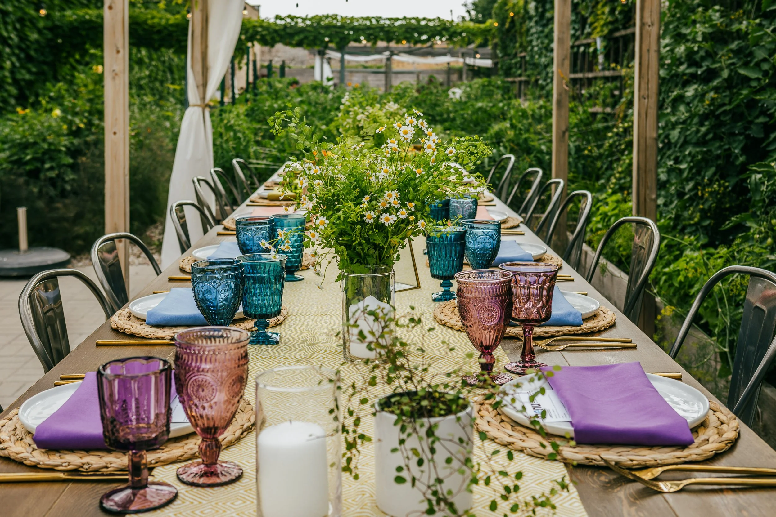 A decorated outdoor dining table with colorful glassware, a centerpiece of daisies, and place settings, surrounded by greenery and a pergola.