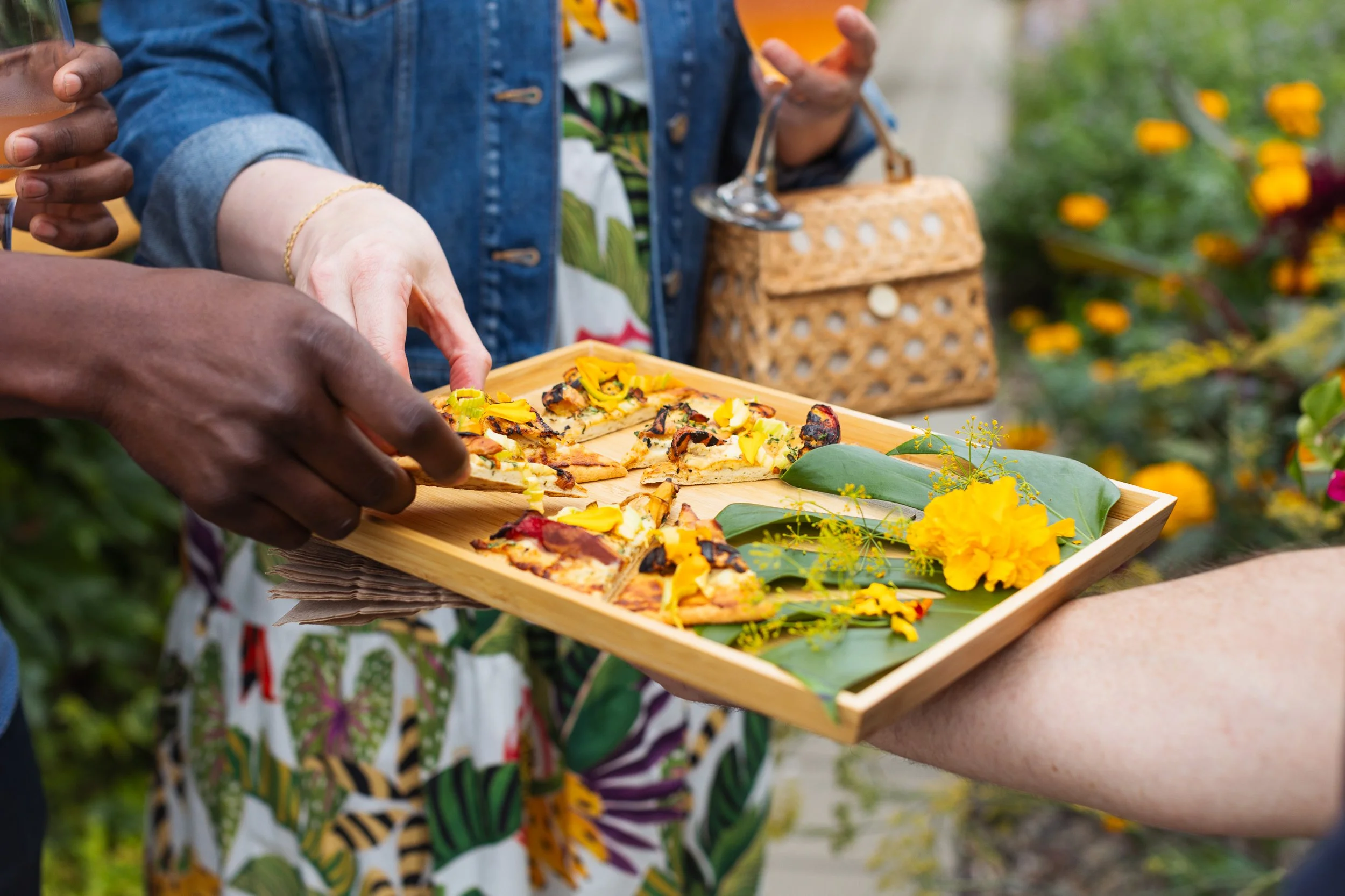 Person in patterned dress holding a wooden tray with slices of pizza topped with yellow flowers, with another person serving. In the background, someone is holding a glass of drink and a woven basket, outdoors surrounded by flowers.