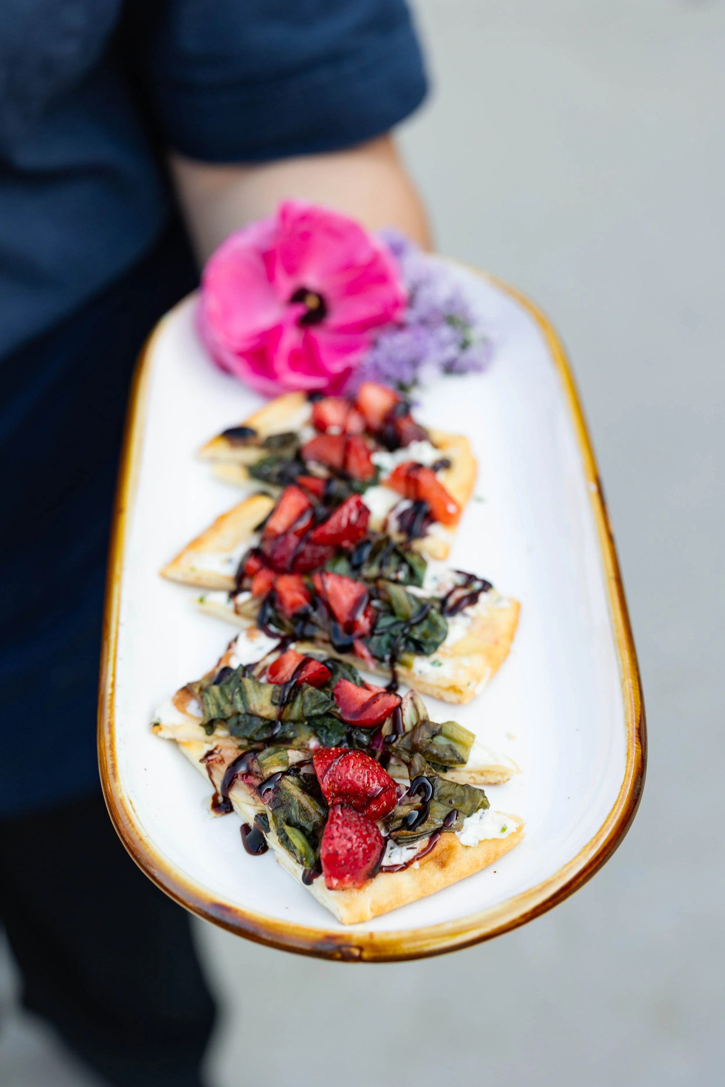 A person holding an oval-shaped plate with five slices of pizza topped with strawberries, strawberries sauce, and basil, garnished with edible flowers and a pink flower on the side.
