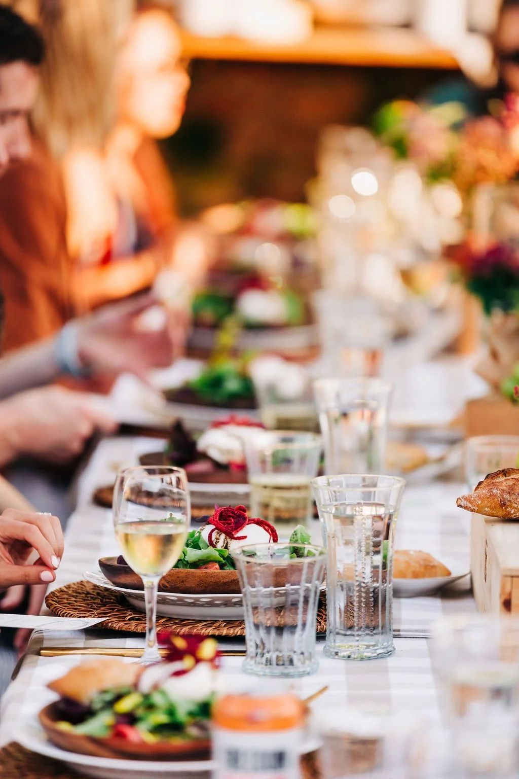 A blurry outdoor dinner party with a long table set with plates, glasses, and food, including a salad with red onions and greens, and various other dishes, with people sitting and eating.
