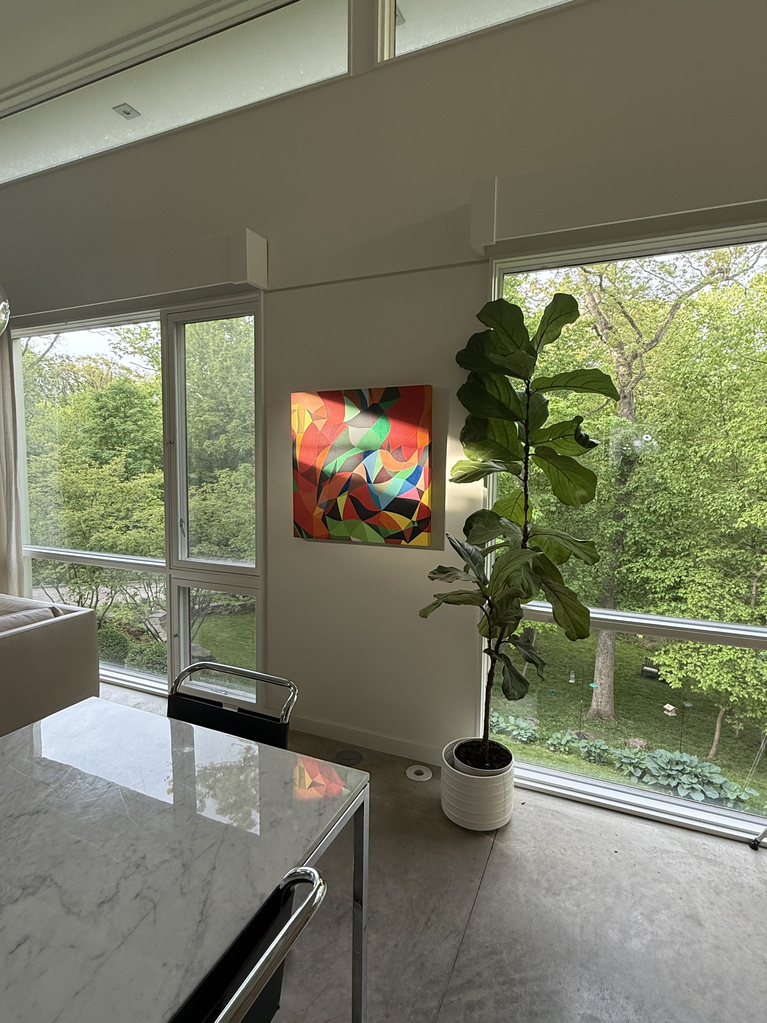 Living room with large windows showing green trees outside, a colorful abstract painting on the wall, a tall potted plant, a marble table, and part of a beige sofa.