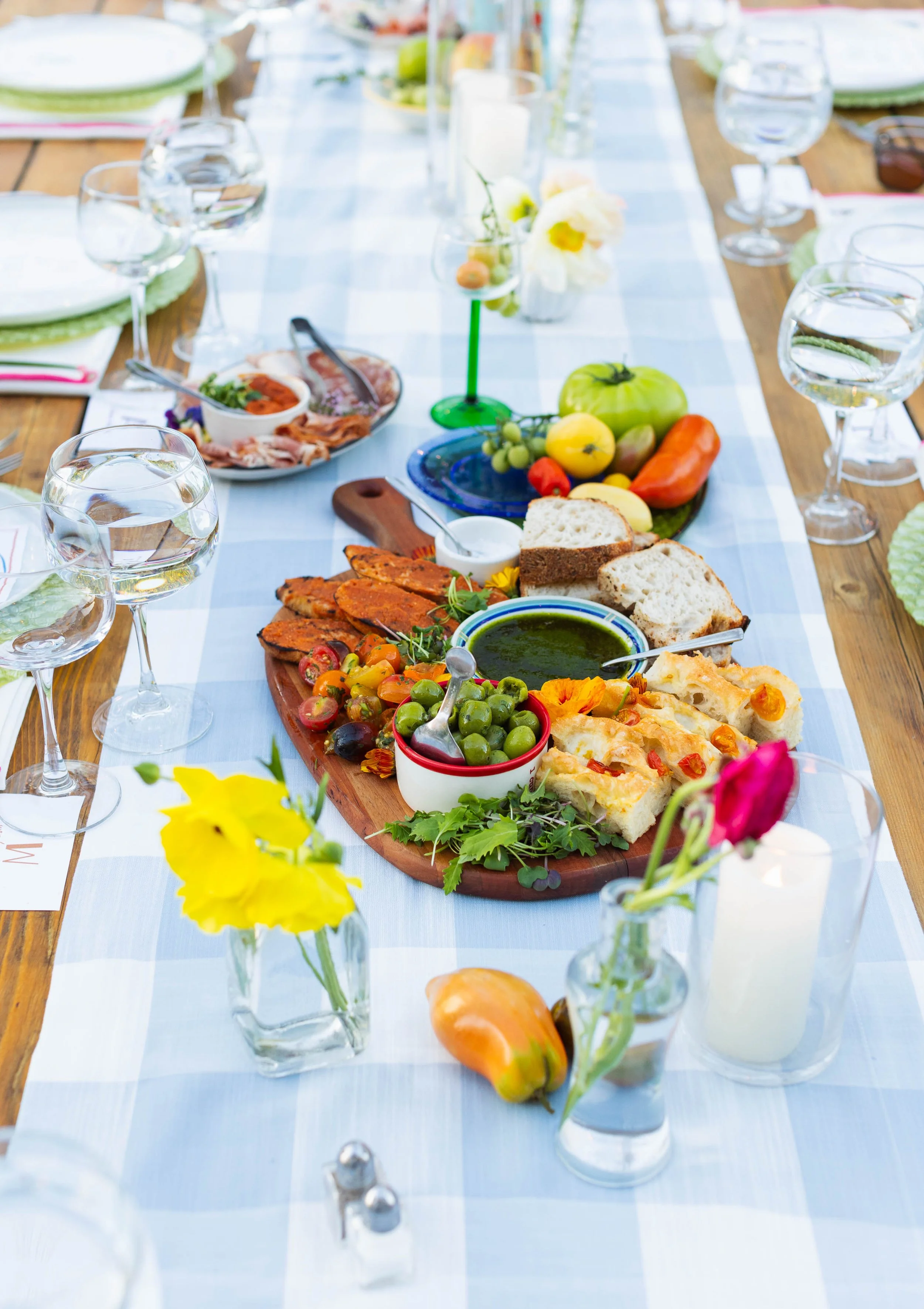A long dining table set for a meal with a white tablecloth, decorated with a flower arrangement and surrounded by empty wine glasses, with a mix of bread, fruit, and food on a wooden serving board and plates.
