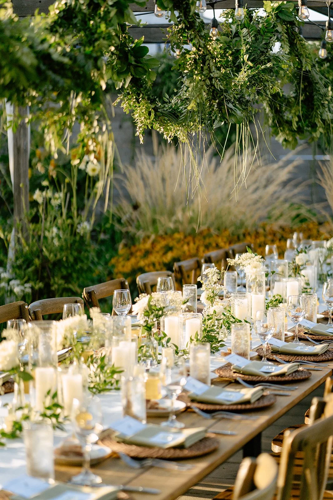An outdoor dining setup with a long wooden table decorated with white flowers, candles, and glassware, beneath string lights and hanging greenery.