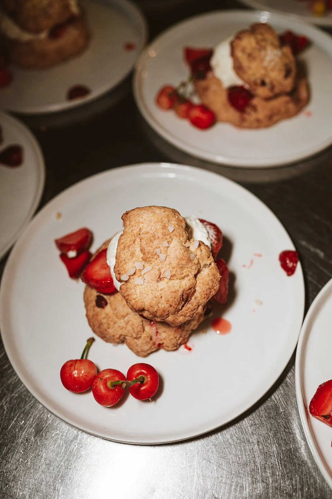 A plate with a large choux pastry topped with sea salt flakes, strawberries, and whipped cream, garnished with fresh cherries, on a white plate. Multiple similar plates are visible in the background.
