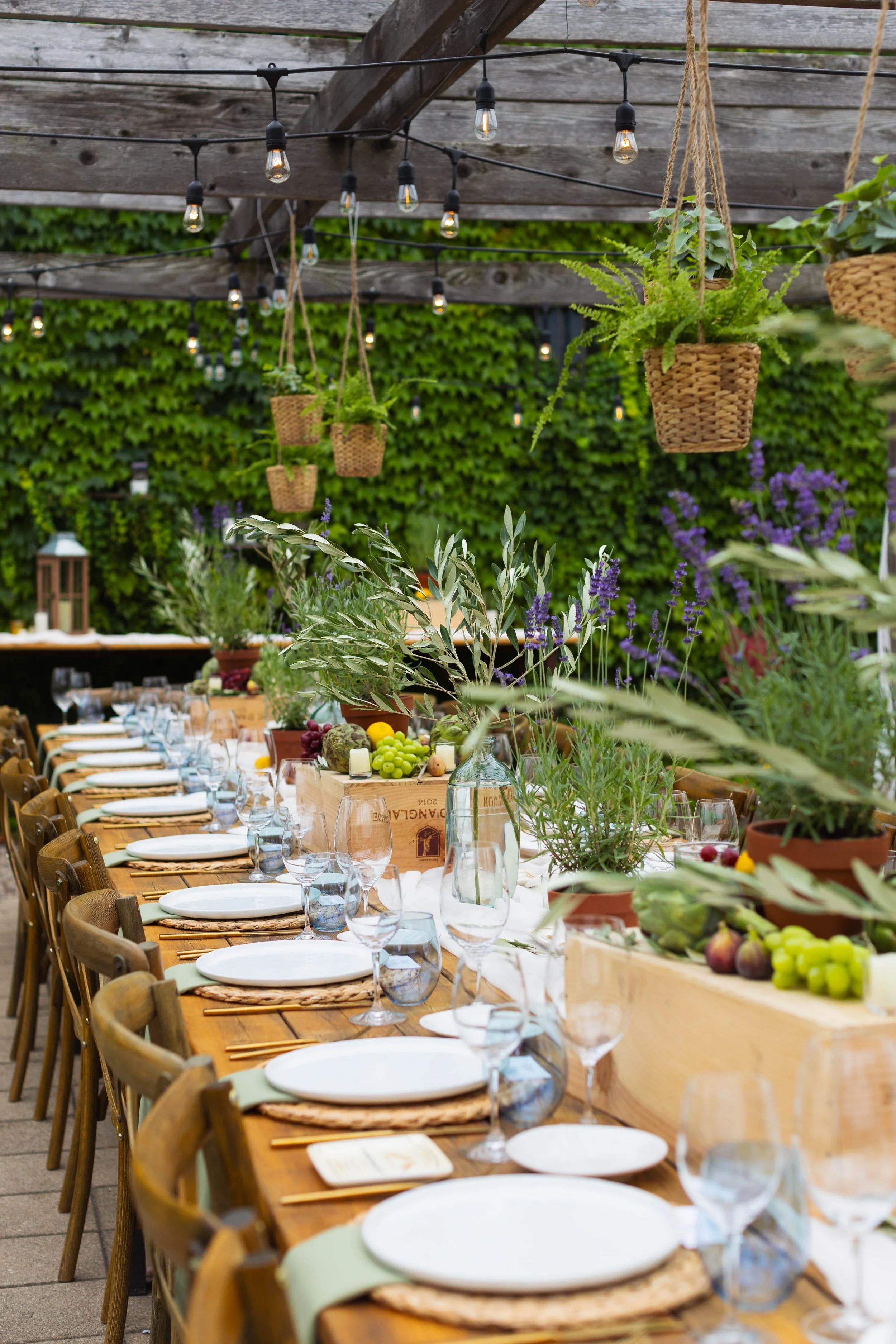 Long wooden banquet table set with white plates, wine glasses, and cutlery, decorated with potted plants, figs, grapes, and lemons. Hanging wicker baskets with green plants are suspended overhead, and string lights are strung above. The setting appea