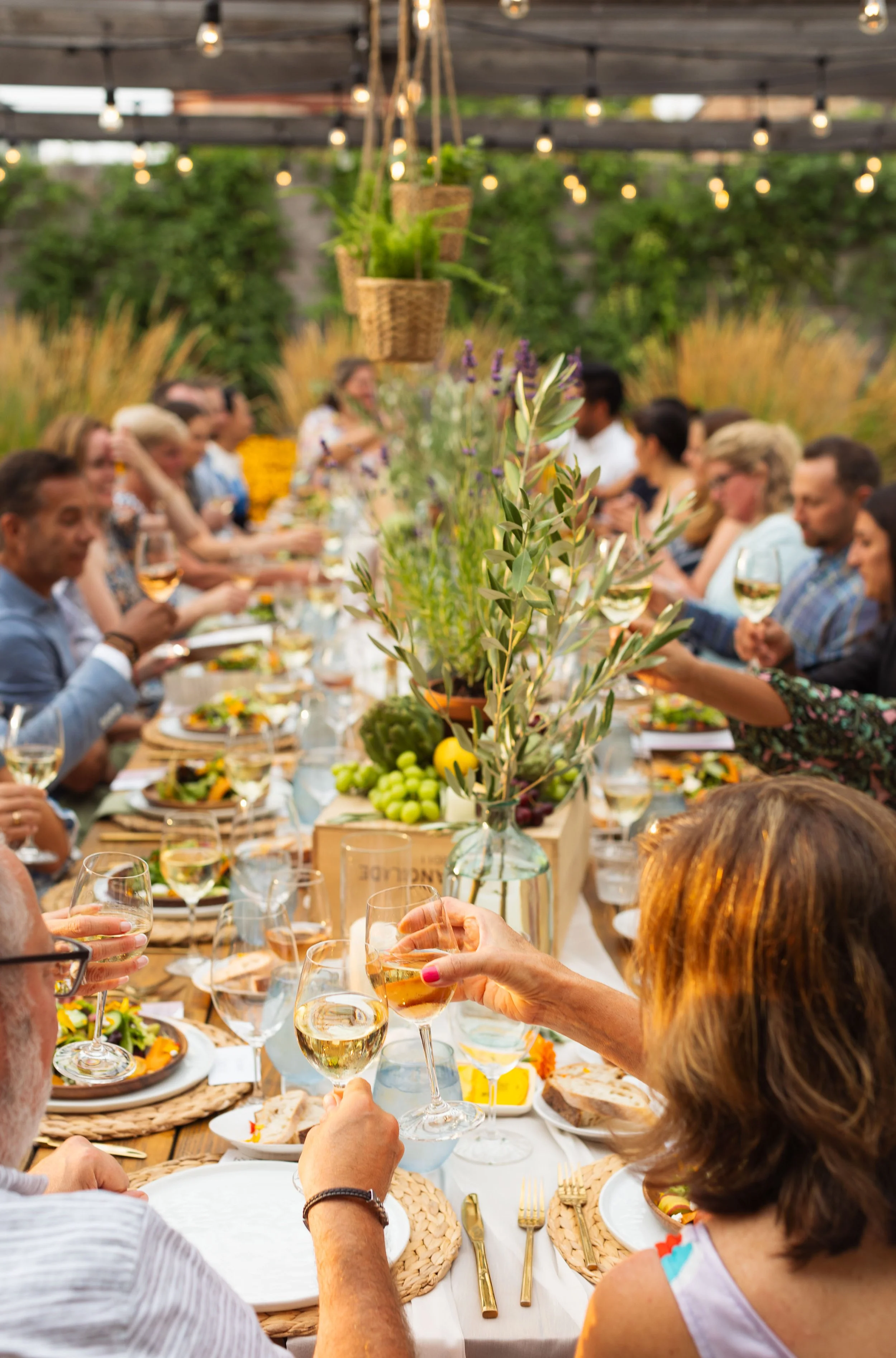 People gathered around a long outdoor table under a pergola with hanging lights, enjoying a meal and drinks, with floral arrangements down the center of the table.