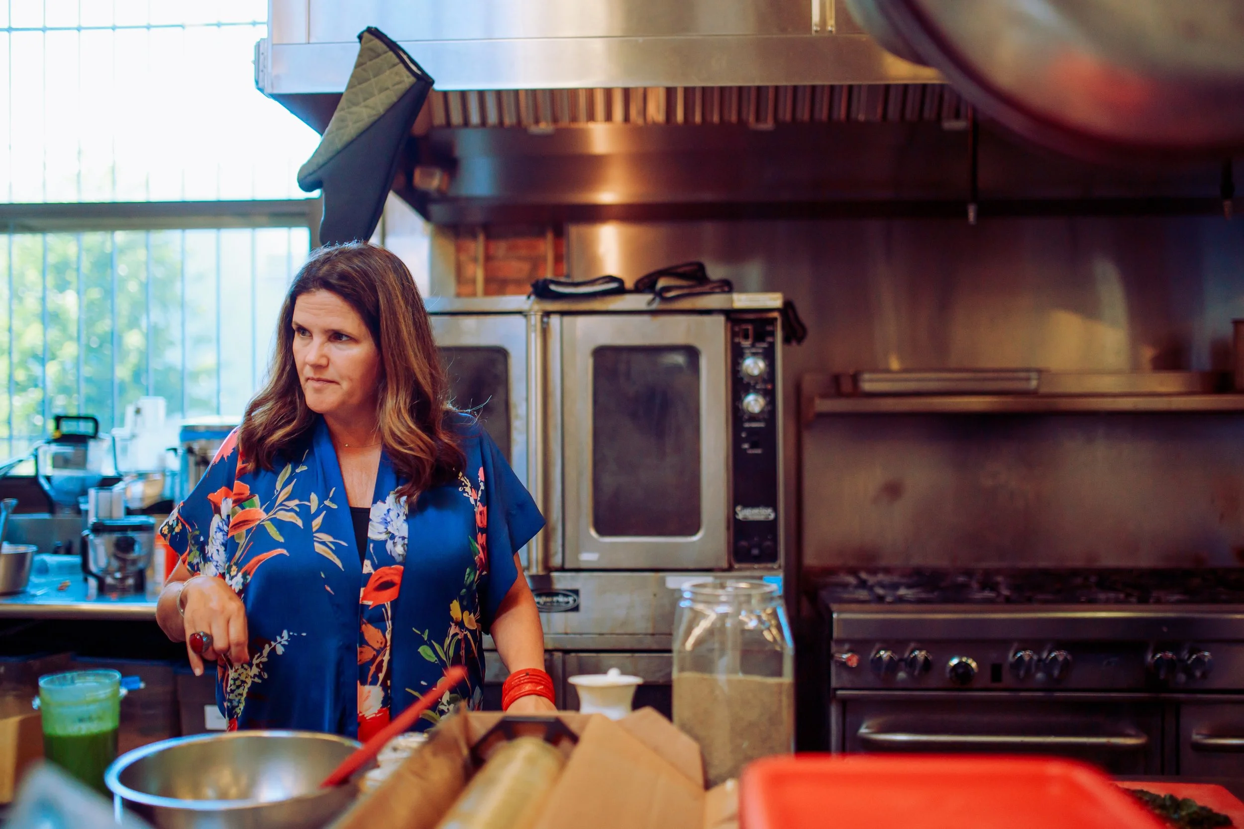 Woman with long brown hair in a blue floral shirt cooking in a commercial kitchen with stainless steel appliances.