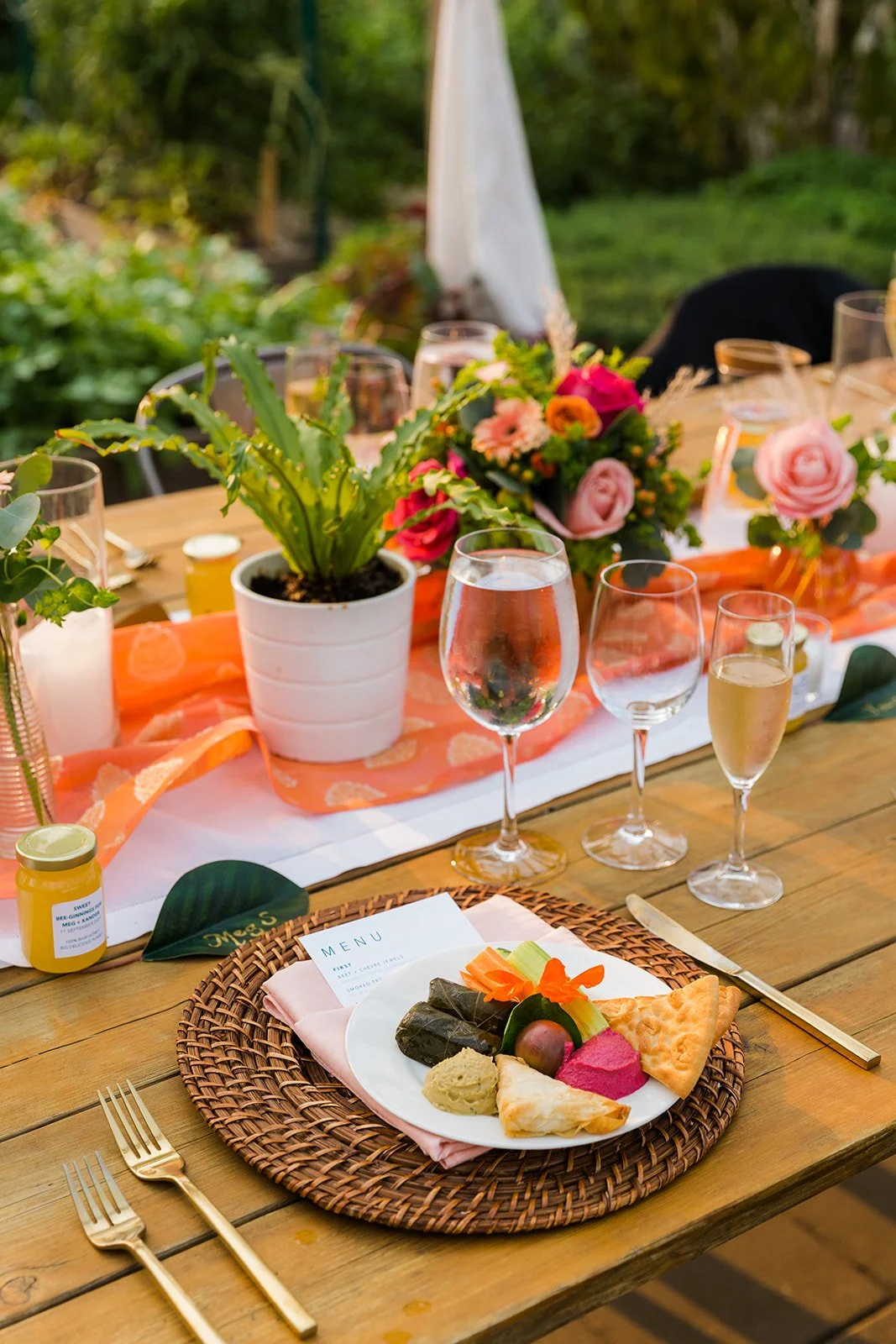 A table set for an outdoor celebration with a centerpiece of colorful flowers, glasses of drinks, a small potted plant, and a plate of assorted foods including rolled wrappers, dip, and crackers.
