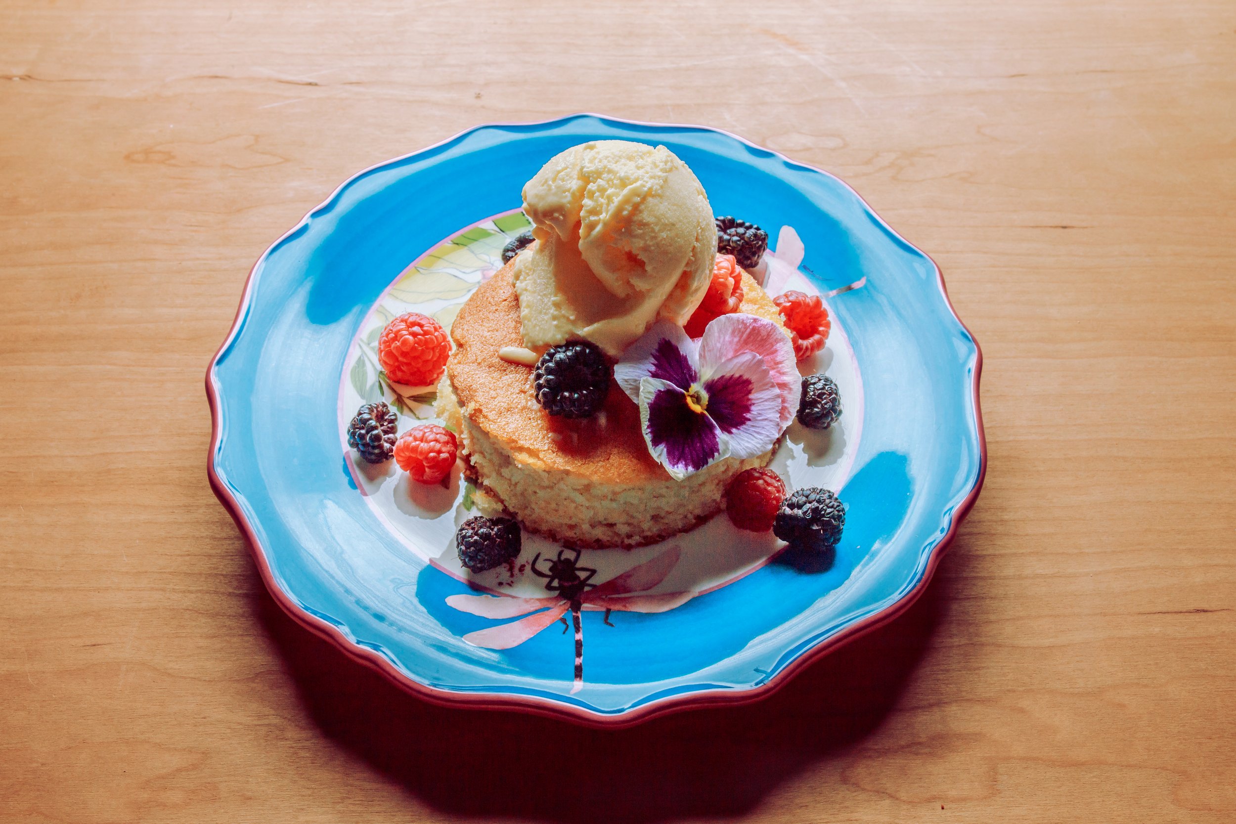A dessert plate with a piece of cake, a scoop of vanilla ice cream, and various berries, garnished with an edible flower, on a decorated ceramic dish placed on a wooden surface.