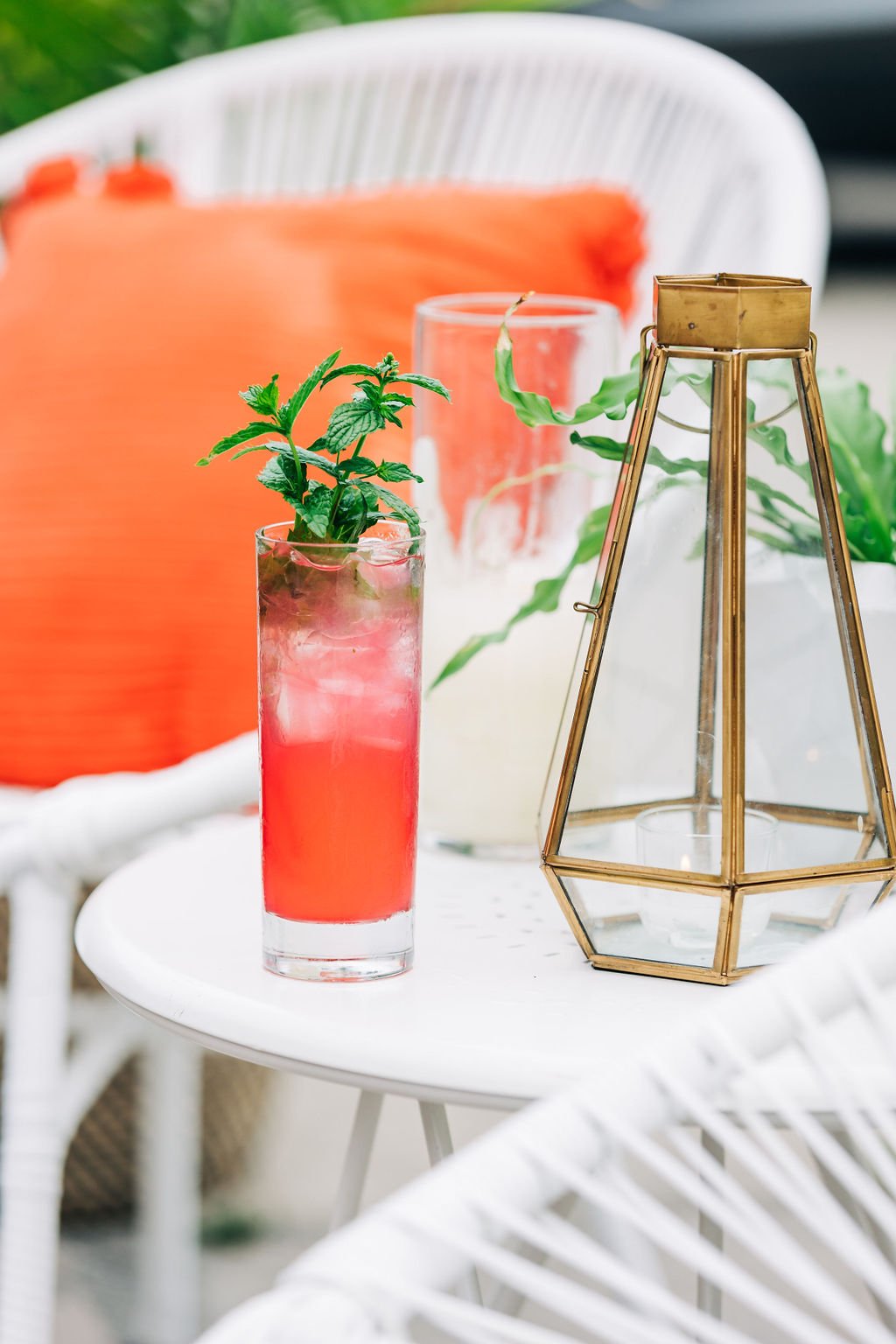A tall glass filled with pink cocktail and ice, garnished with fresh mint leaves, is on a white round table. Behind it, there are clear glass containers and a geometric gold lantern, with a white chair, orange cushion, and green plants in the backgro