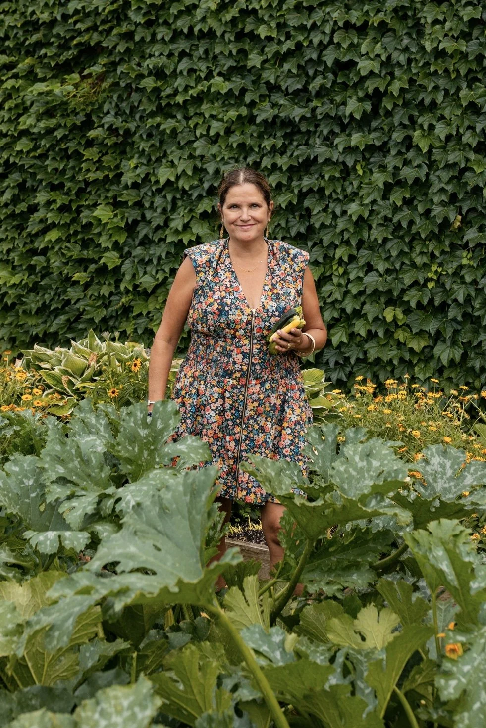 A woman wearing a floral dress is standing in a garden filled with green leafy plants and yellow flowers, holding some vegetables.