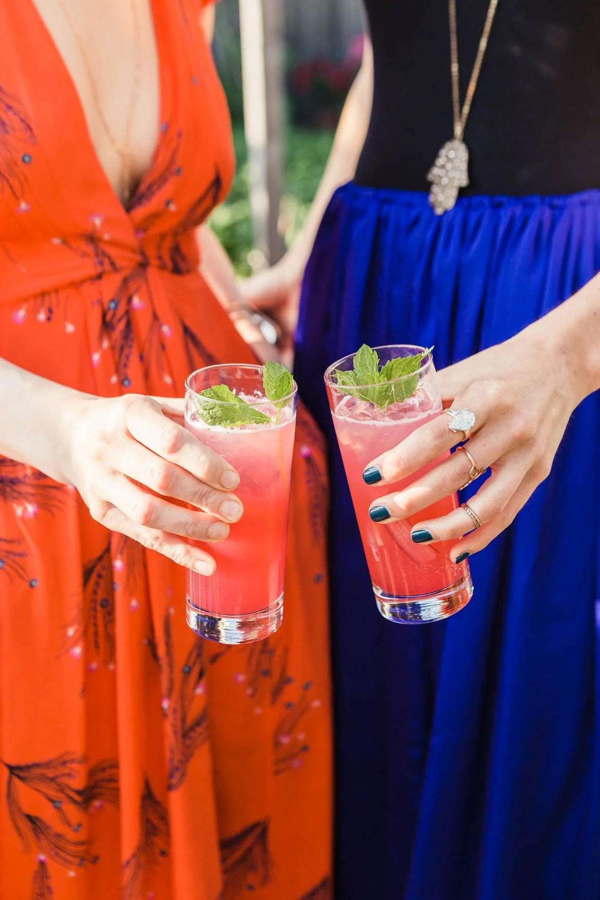 Two women holding pink drinks garnished with mint leaves at an outdoor gathering.