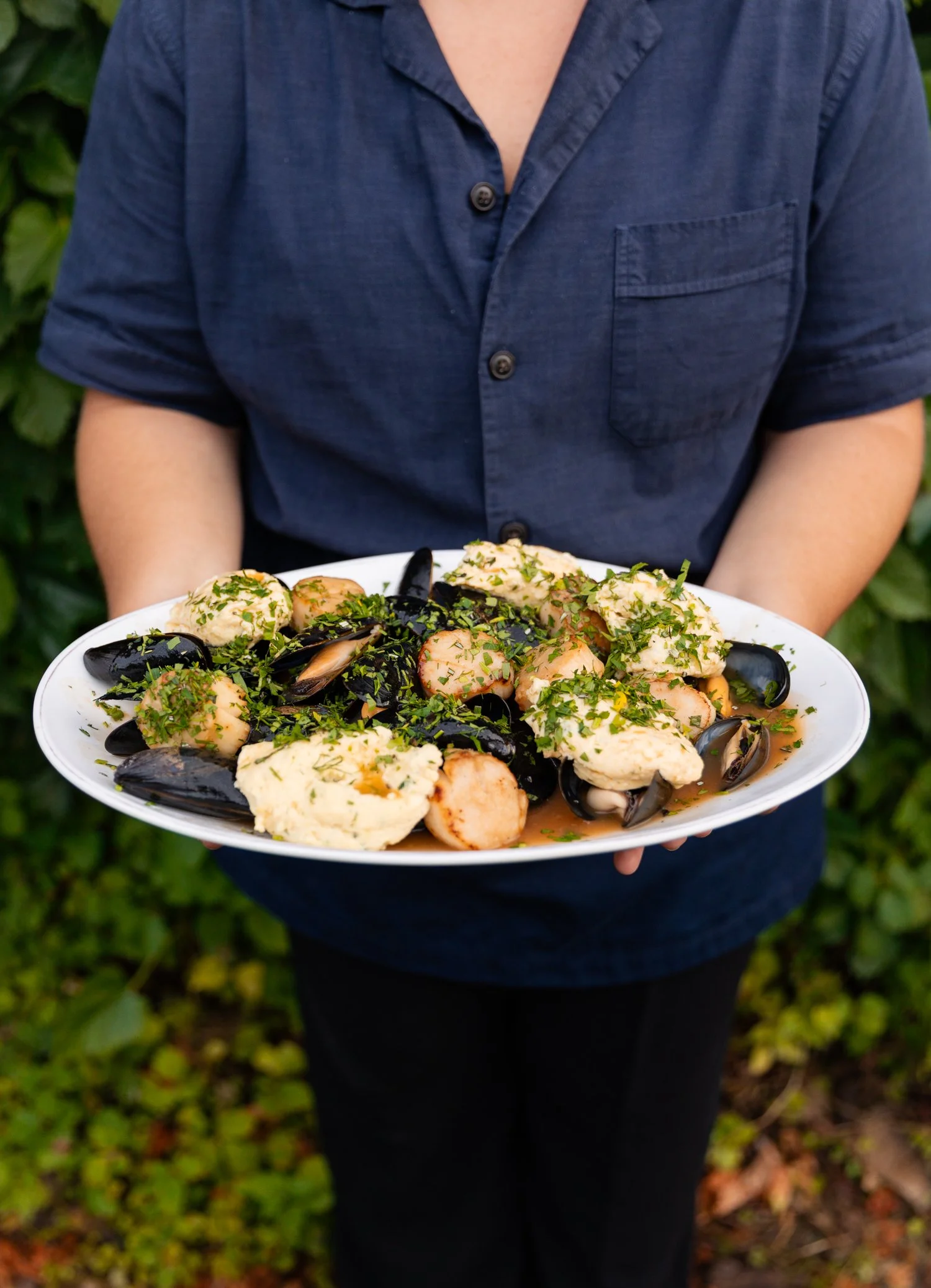 Person holding a large white platter of seafood, including mussels, shrimp, and fish, garnished with herbs, outdoors with green foliage background.