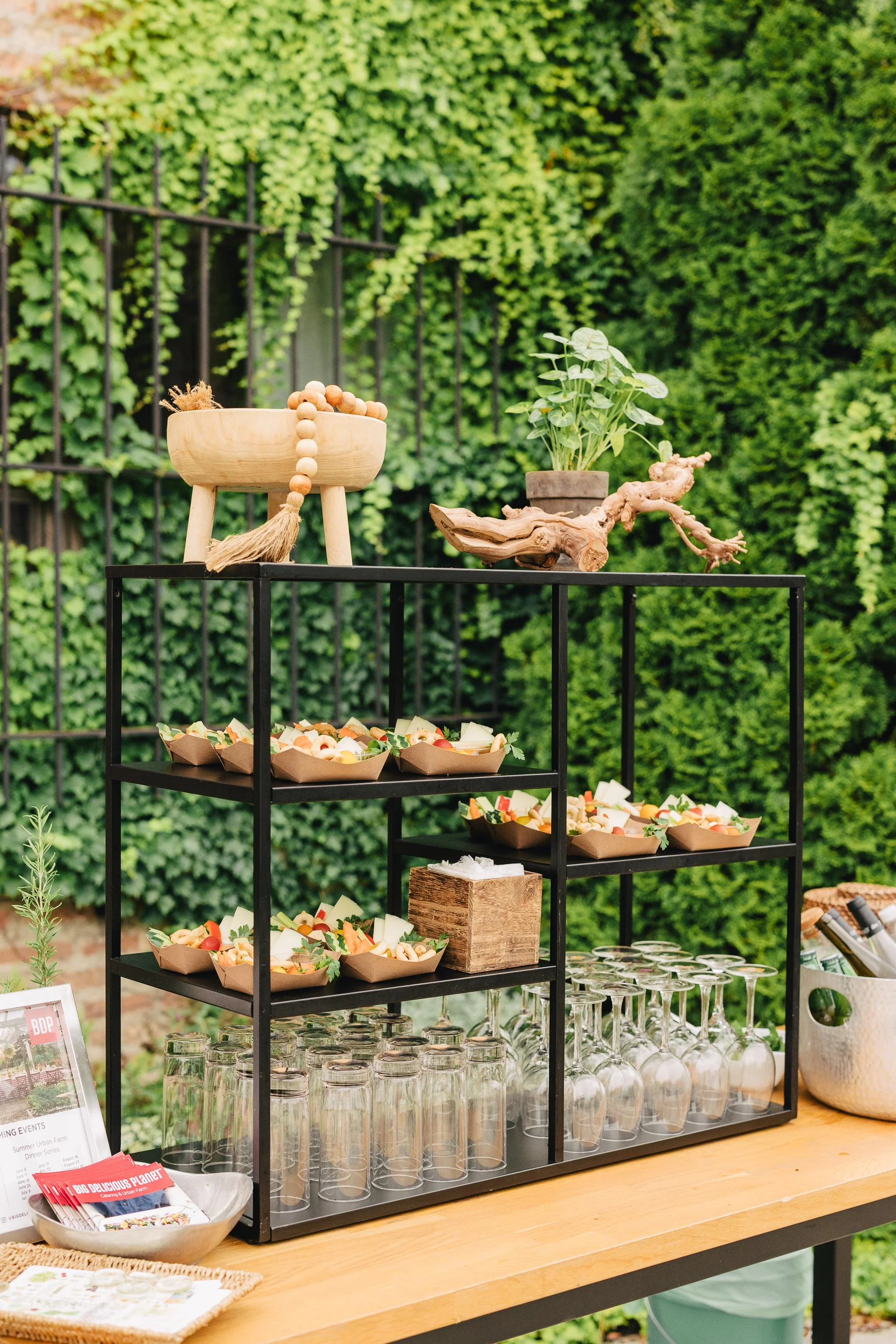 Display of small food bowls, empty glasses, and decorative items on a black metal stand at an outdoor event with lush green hedge in the background.