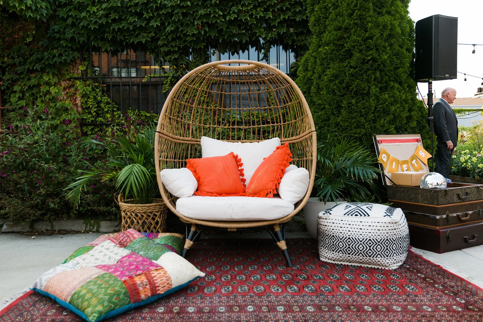 Outdoor seating area with a wicker hanging chair, white and orange cushions, colorful pillows, a patterned pouf, a patchwork quilt, potted plants, a decorative box with 'GIFTS' written on it, and a man in a suit standing near a speaker and lights.