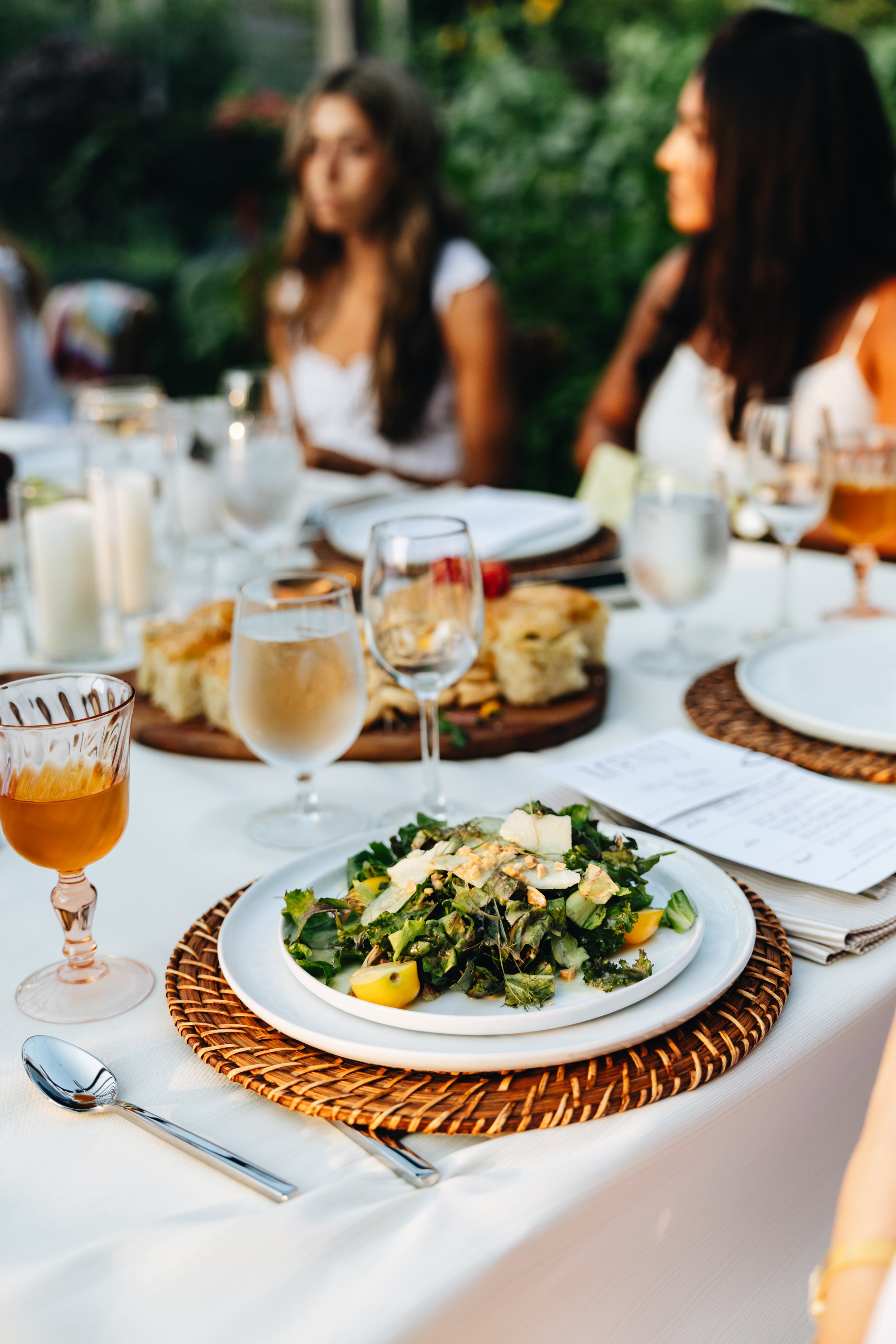 A table set for a meal outdoors with a salad, glasses of drinks, and a menu, and blurred guests in the background.