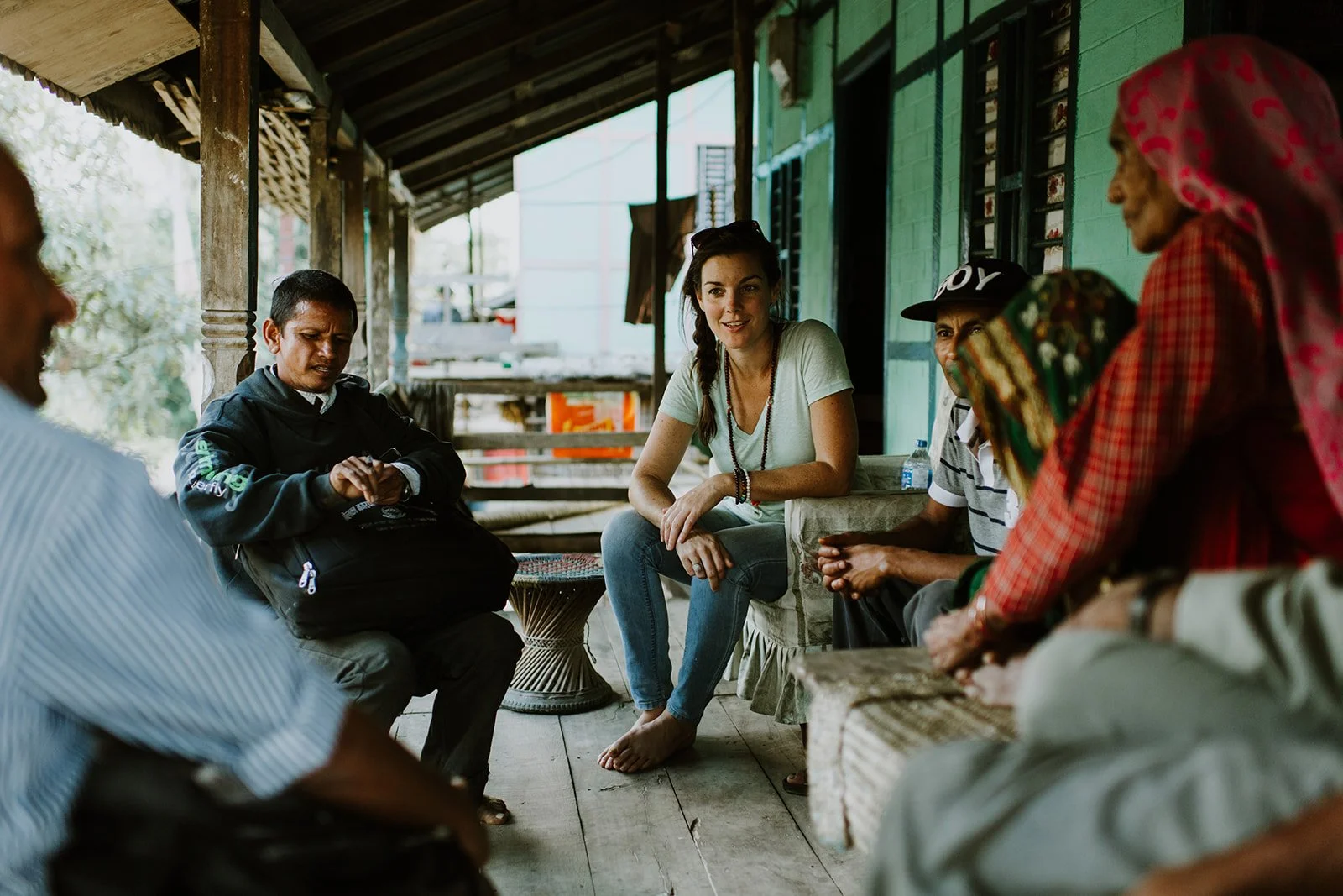 A group of people sitting in a circle on a porch, engaging in conversation; the porch has wooden floors and a roof, with trees and a distant building in the background.