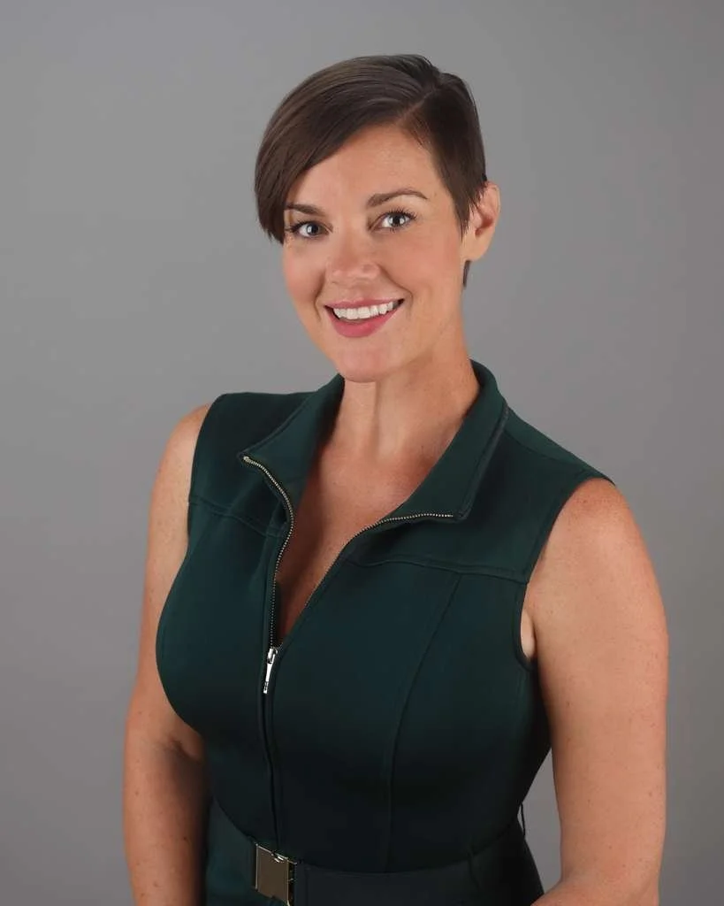 A woman with short brown hair smiling, wearing a sleeveless dark green zip-up top against a plain gray background.