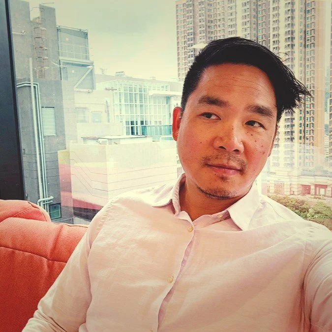 A man with black hair, light skin, and a slight beard, wearing a light-colored shirt, sitting on an orange sofa with a cityscape view of tall buildings through a large window behind him.