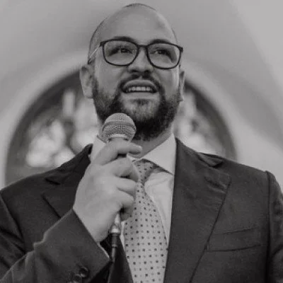 A man with glasses, a beard, and a mustache holding a microphone, speaking at a formal event in a church or cathedral setting.