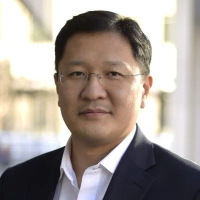 Professional portrait of an Asian man in a suit and glasses, indoors with a blurred background.