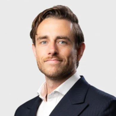 A man with dark hair and a beard wearing a dark suit jacket and white shirt, smiling slightly against a light gray background.