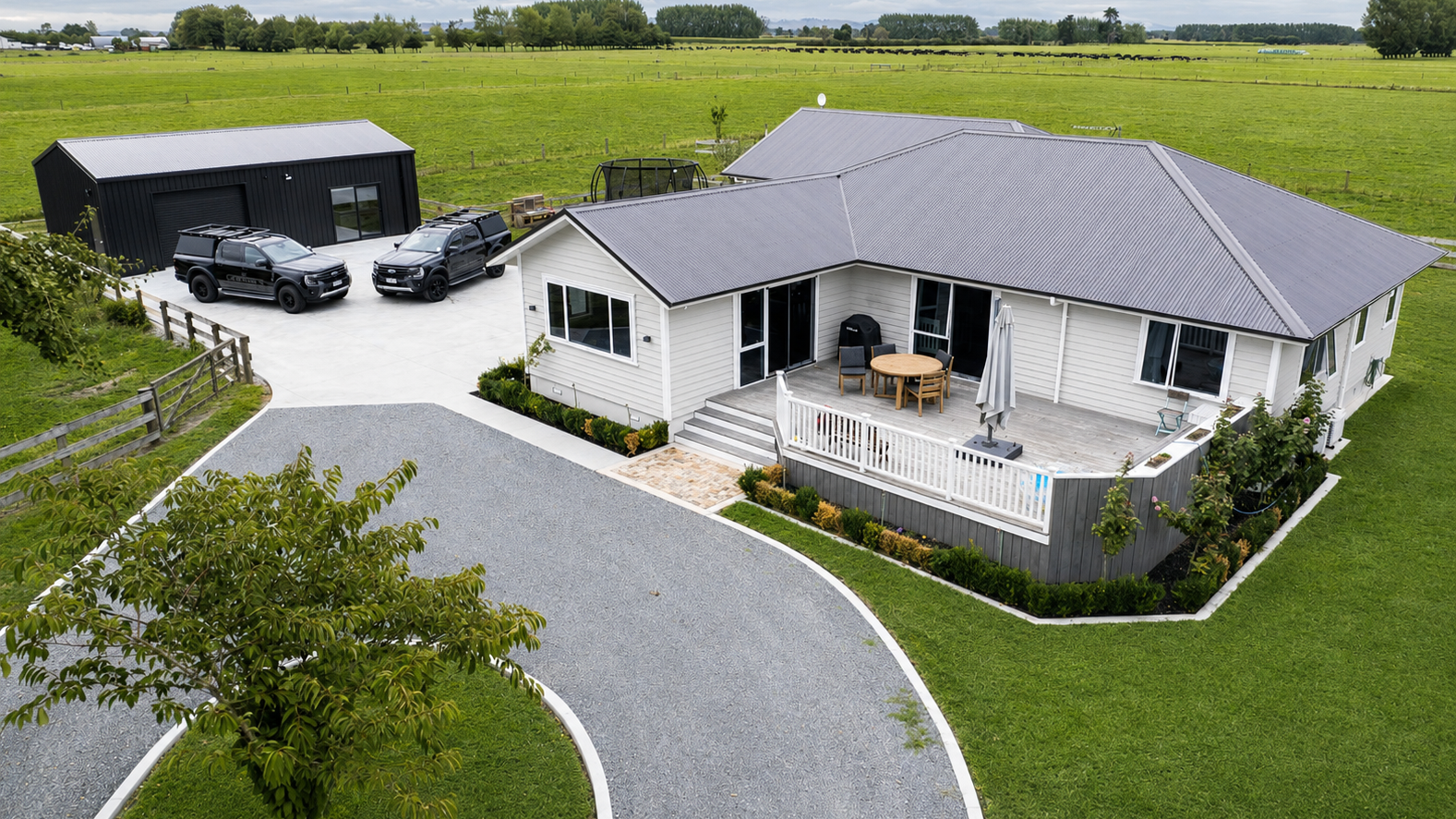 A modern house with a spacious porch and a large backyard, situated in a rural area with green fields and trees in the background. The house has a gray metal roof and white siding, with outdoor furniture on the porch. There are two black vehicles parked on a concrete driveway, along with a black storage building nearby.