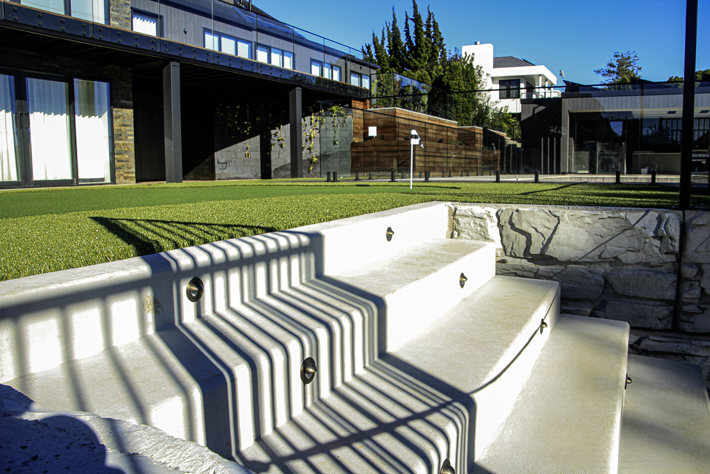 Modern house with glass walls, green grass, and outdoor concrete stairs with shadows from a metal railing.