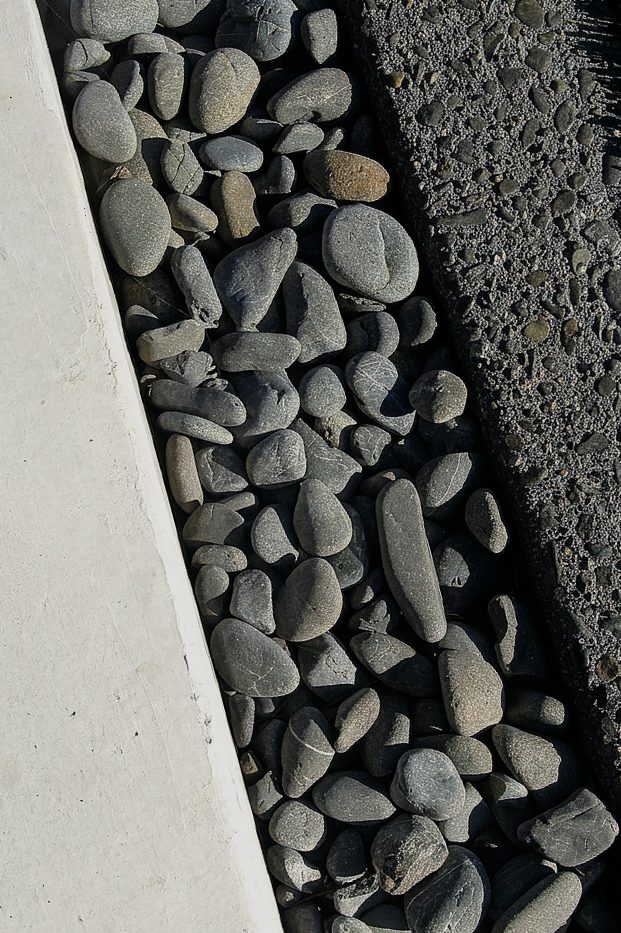 Close-up of gray rounded pebbles and rocks on a beach, with a concrete barrier on the left and a dark rocky area on the right.