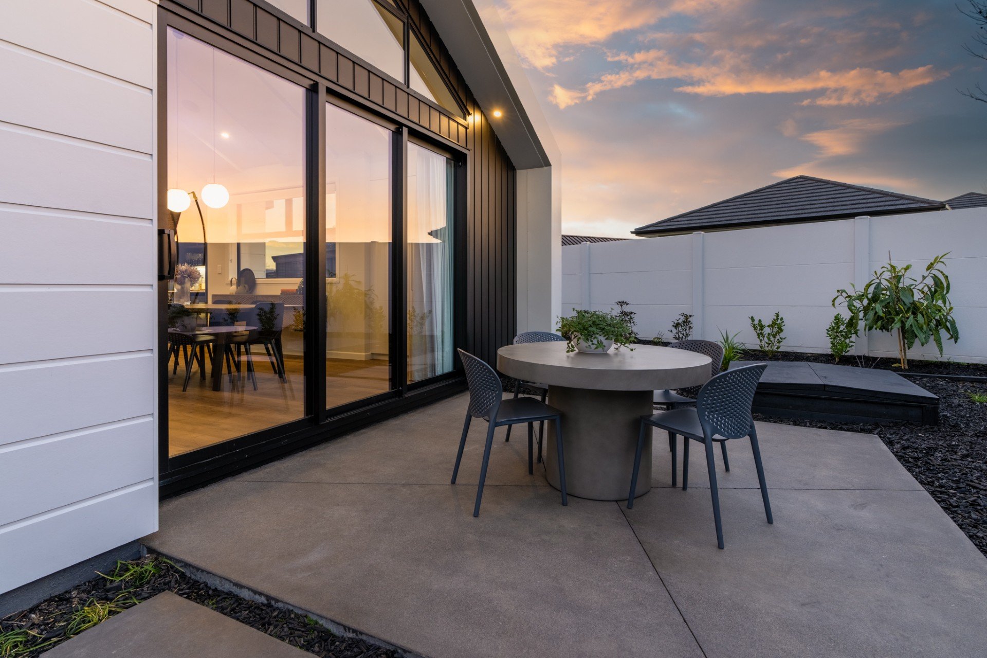 Modern house patio at sunset with a round table and four chairs, small garden with plants, and large sliding glass doors leading inside.