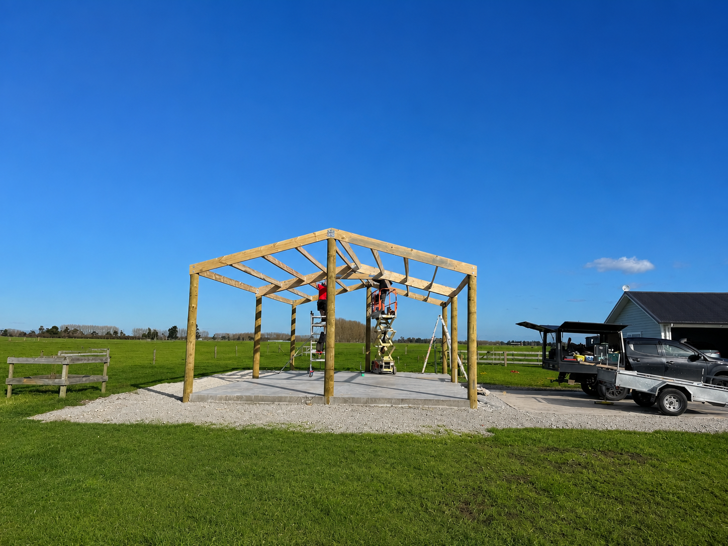Construction site with a wooden structure frame, workers on ladders, and a truck parked nearby on a rural field under a blue sky.