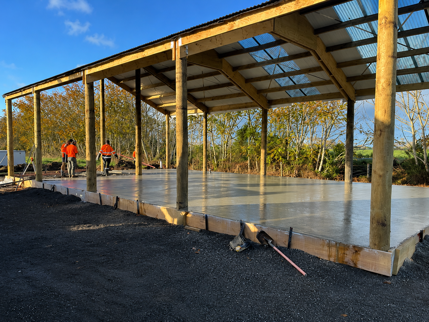 Construction workers pouring a concrete slab under a wooden and metal roof structure on a sunny day, trees in the background.