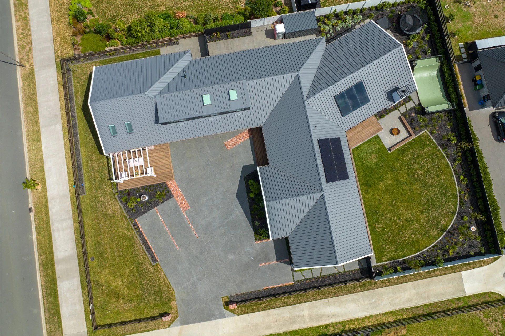 Aerial view of a modern house with a metal roof, solar panels, a backyard with a patio, a trampoline, and a small swimming pool, surrounded by a lawn and garden, with a street and sidewalk nearby.