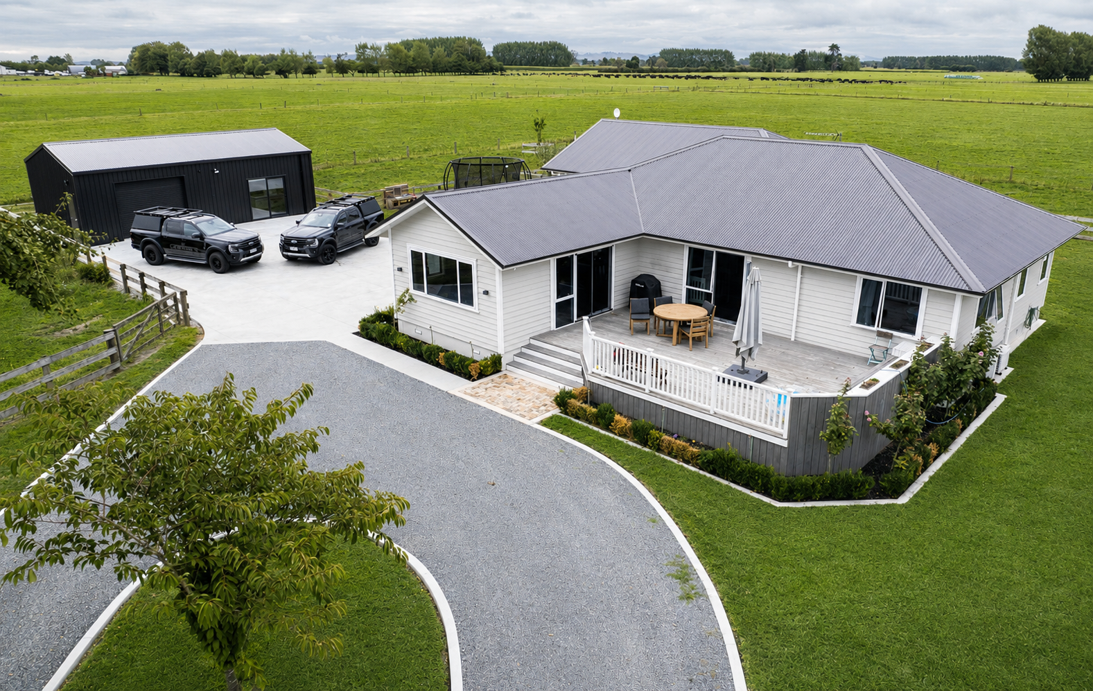 A white single-story house with a gray metal roof, a wooden deck with outdoor furniture and a closed umbrella, a driveway with gravel and pavement, three black vehicles, a black shed, and a green landscape with trees and farmland in the background.