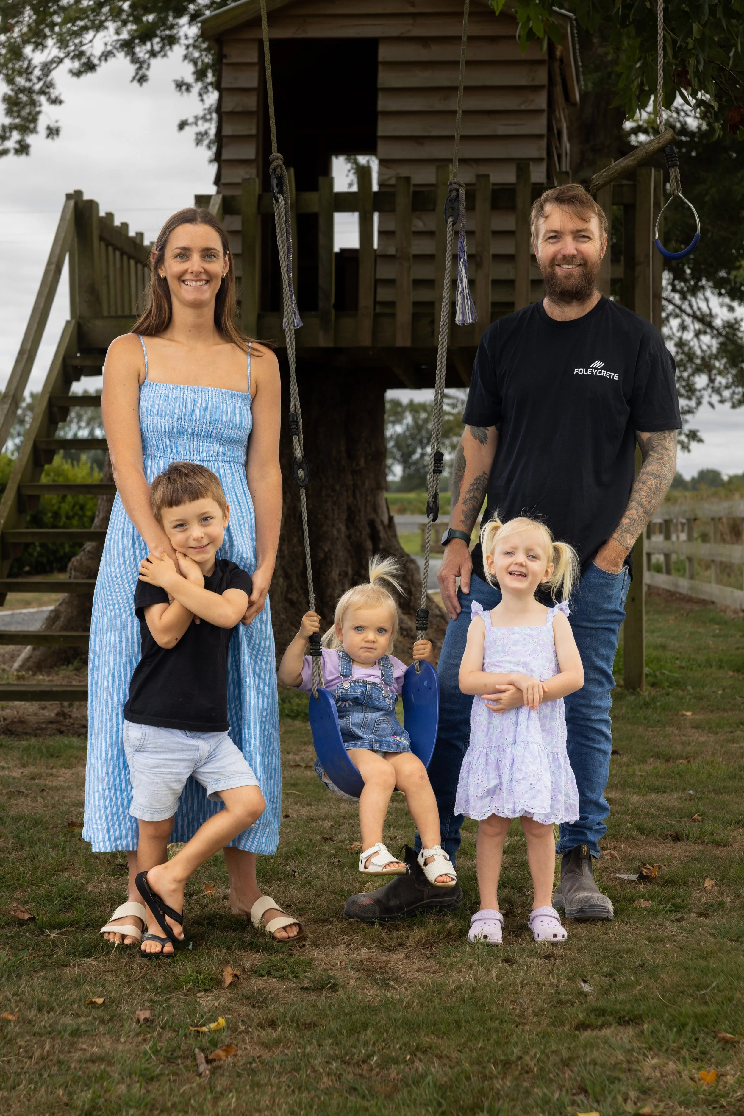 Family posing outdoors in front of a treehouse, with two adults and four children. The woman in a blue dress and the man in a black t-shirt stand on either side of the children. One child is seated on a swing, and the other three children are standing, with some holding hands or touching each other.