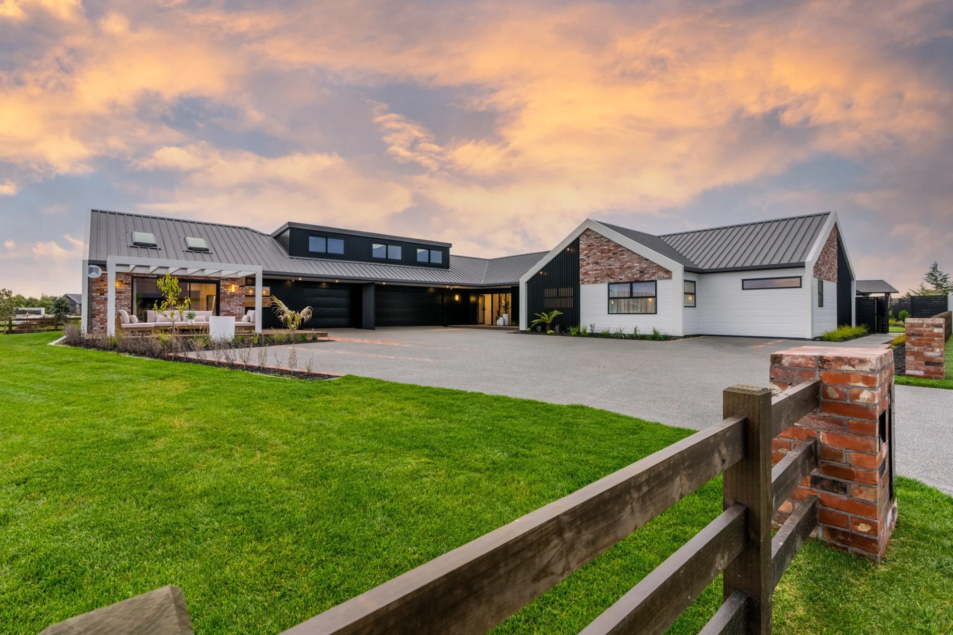 Modern house with black and white exterior, brick accents, and metal roof, surrounded by a green lawn and a wooden fence, under a colorful sky at dusk.