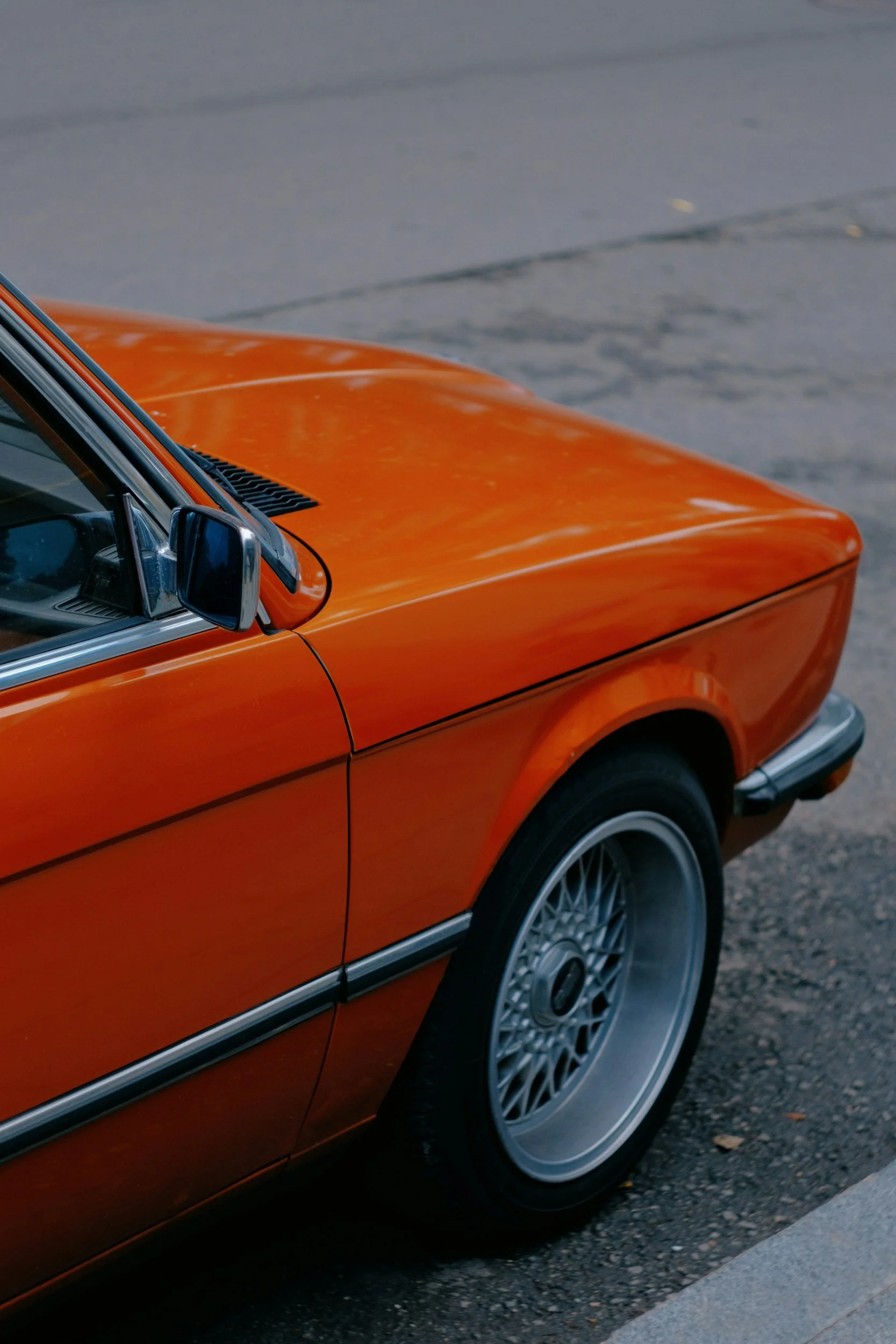Close-up of the front side of an orange vintage car parked on the street, showing part of the door, side mirror, and wheel.