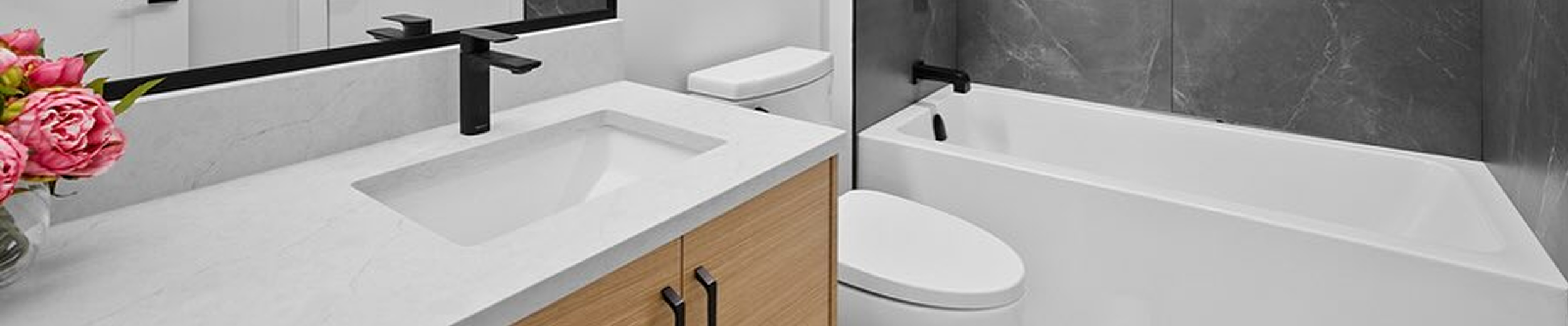Modern bathroom with black fixtures, wooden cabinet, white sink, toilet, and bathtub with dark gray wall tiles, and a bouquet of pink flowers on the counter.