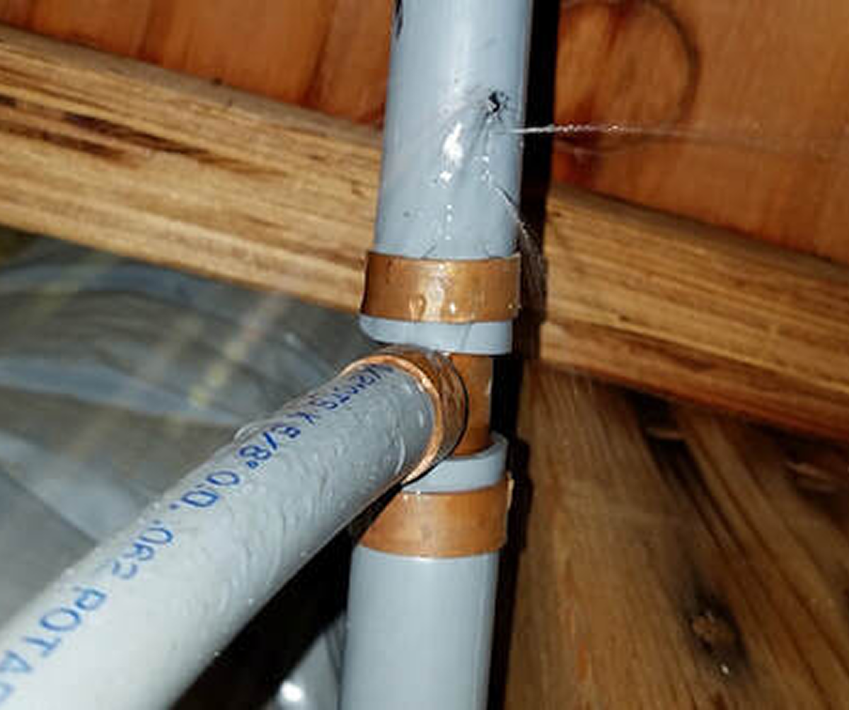 Close-up view of PVC pipes connected with copper-colored couplings, mounted on a wooden wall. Visible spider web on the pipes.