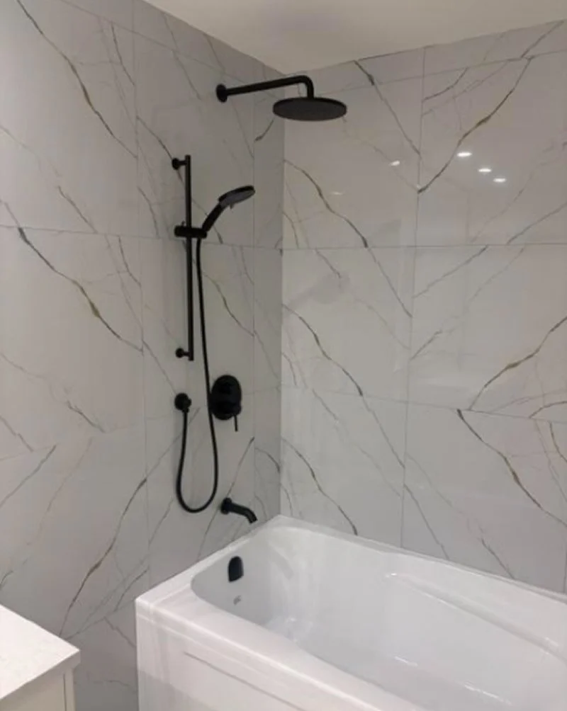 A modern bathroom shower and bathtub with large white marble tiles with gray veining, black shower fixtures, a rainfall showerhead, and a hand shower.