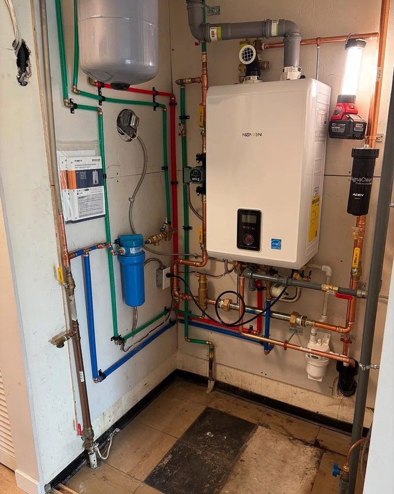 A plumbing and heating system with copper, plastic, and flexible pipes, a large white tankless water heater, a blue filter or pressure tank, and various valves and gauges installed on a wall in a utility closet.