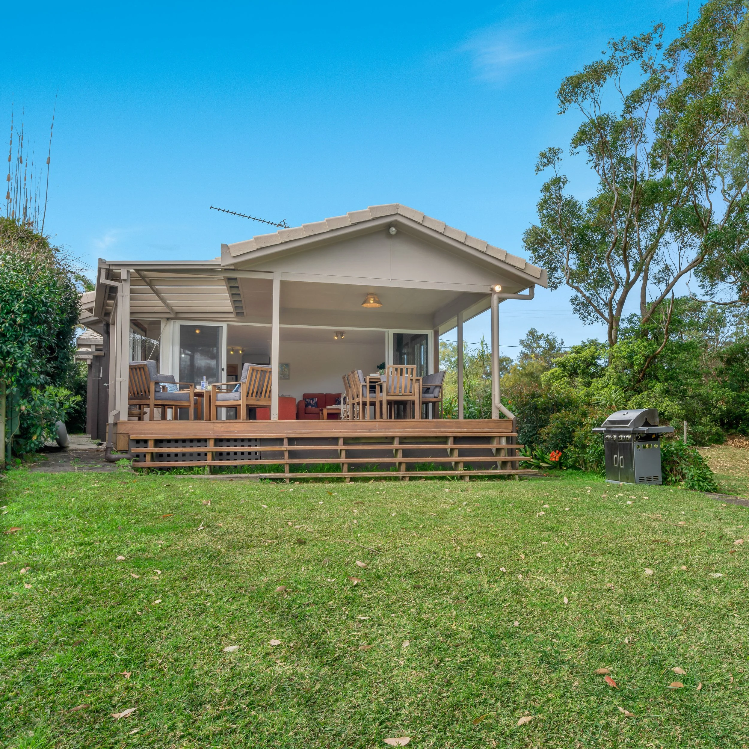 Backyard patio area with a wooden deck, outdoor furniture including chairs and a sofa with cushions, a barbecue grill, and a covered roof, surrounded by green bushes and trees under a clear blue sky.