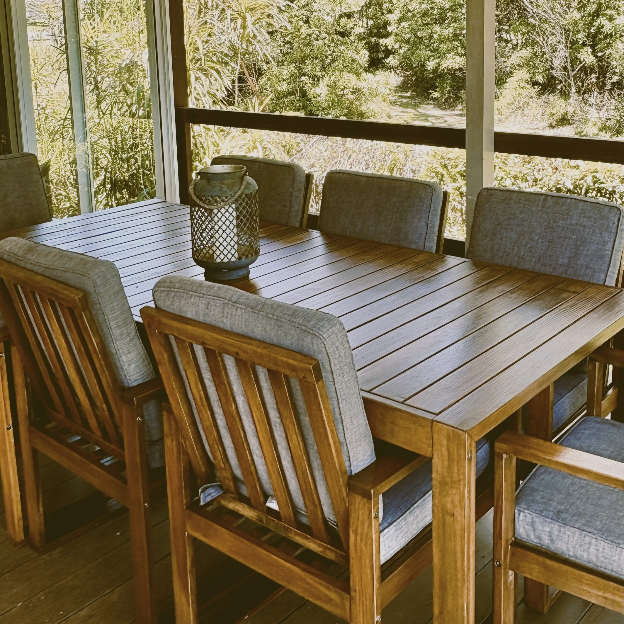 Wooden outdoor dining table with chairs on a porch, with a scenic view of trees and foliage outside.