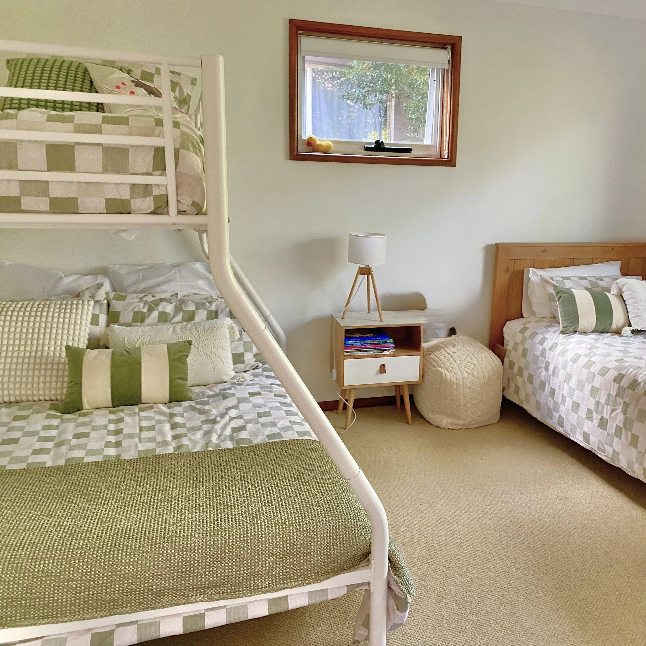 A bedroom with a white bunk bed with green and beige checkered bedding, a wooden nightstand with shelves and books, a white lamp, a window with a wooden frame showing greenery outside, and a green carpet on a beige wall-to-wall carpet.