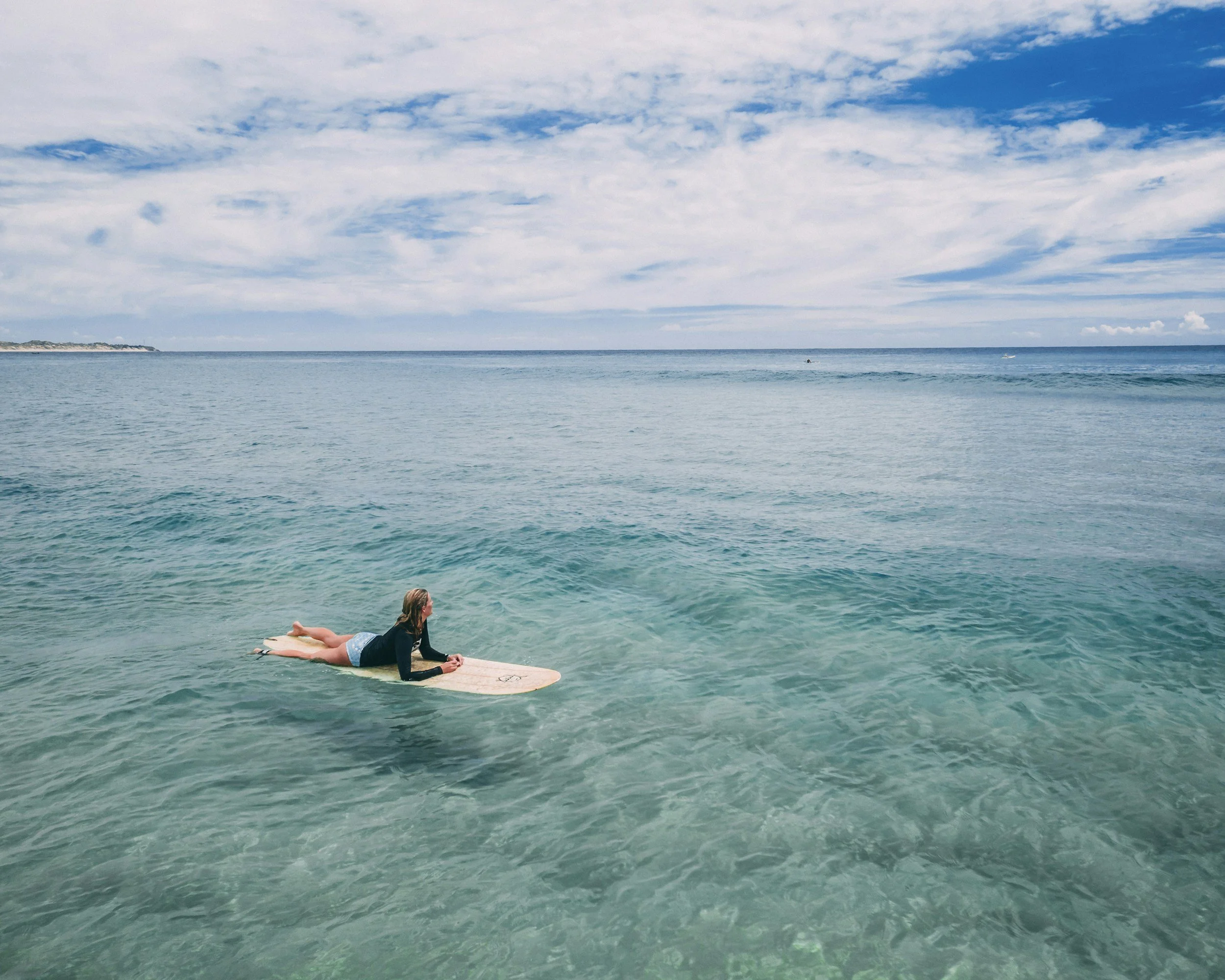Person lying on a surfboard in the ocean, with blue sky and clouds overhead.