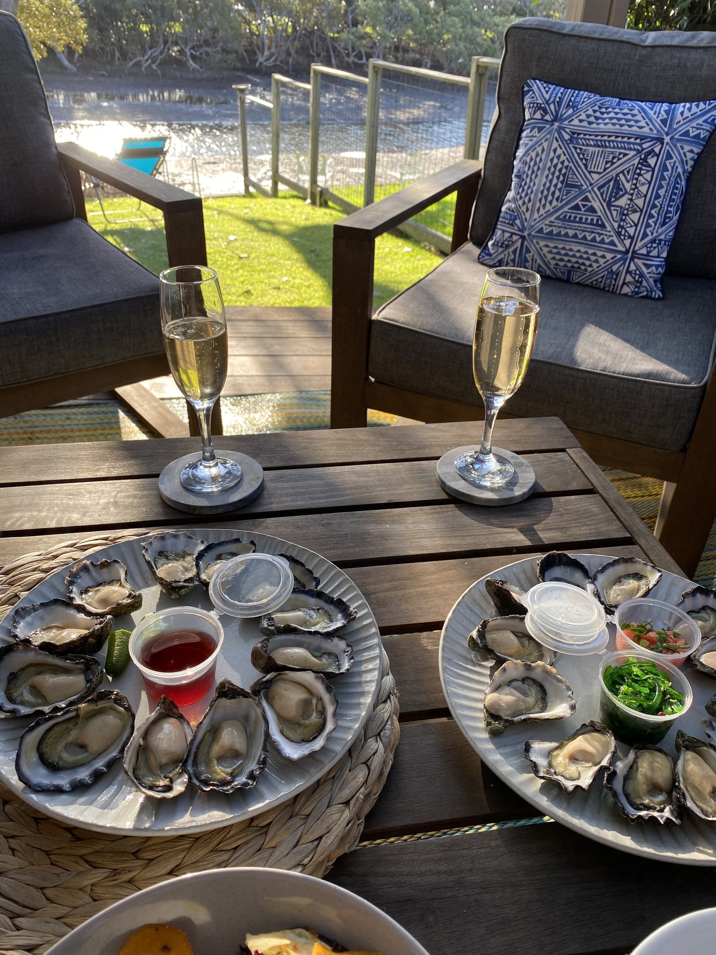 A table set outdoors with plates of fresh oysters, garnishes, and drinks, overlooking the tidal creek with sunlight reflecting off the water.